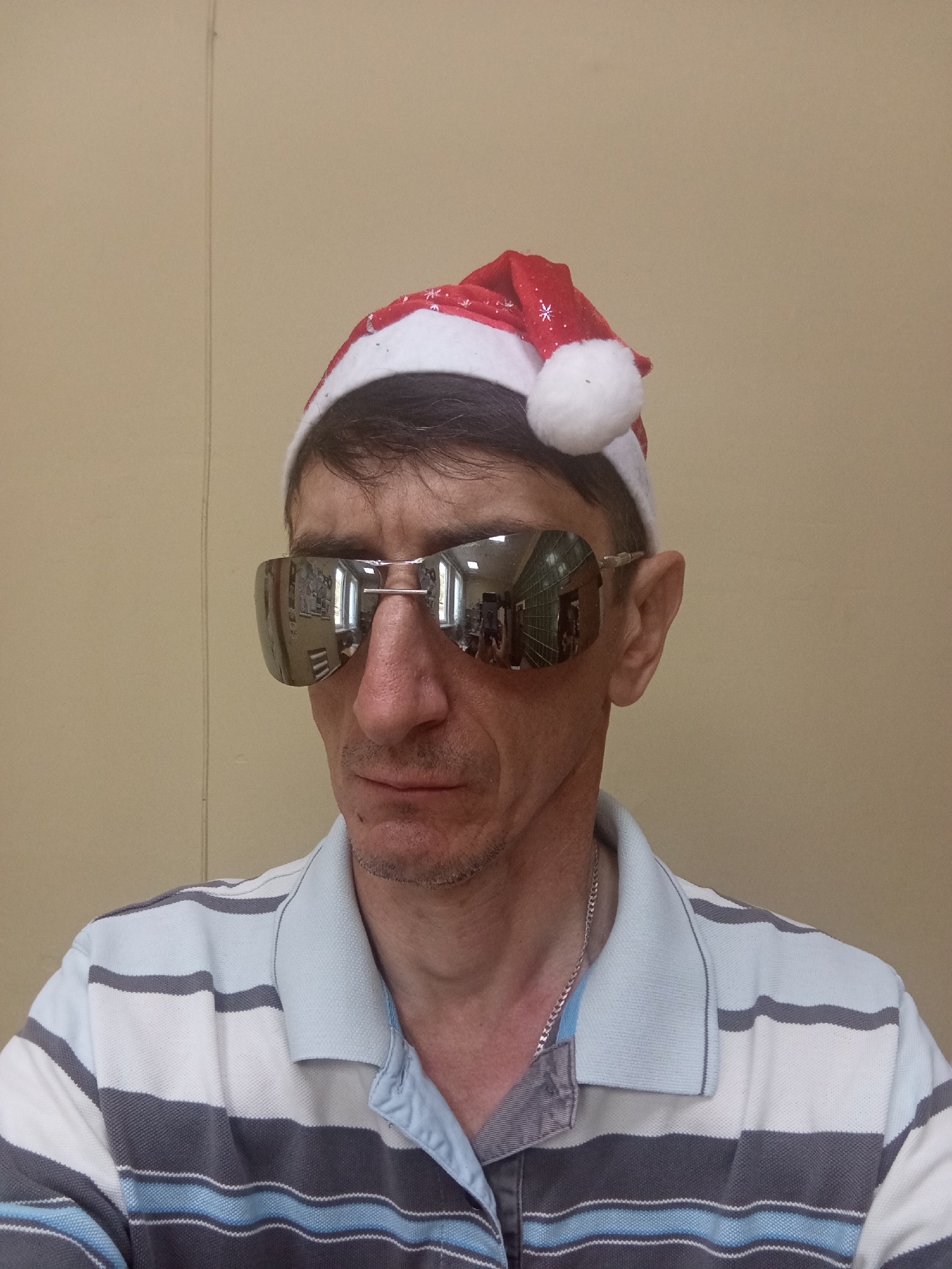 A middle-aged Caucasian man wearing a red and white Santa hat, mirrored sunglasses, and a horizontally striped polo shirt stands in front of a plain, beige wall. The background appears to be an indoor setting with a subtle vertical seam in the wall. The man's expression is neutral, and his face is partially obscured by the large sunglasses, which reflect images of what appears to be a hallway with windows and possibly shelving. The man is also wearing a thin silver chain around his neck. The image does not provide any information to determine a specific location within Крыўлянскі сельскі Савет, Belarus.