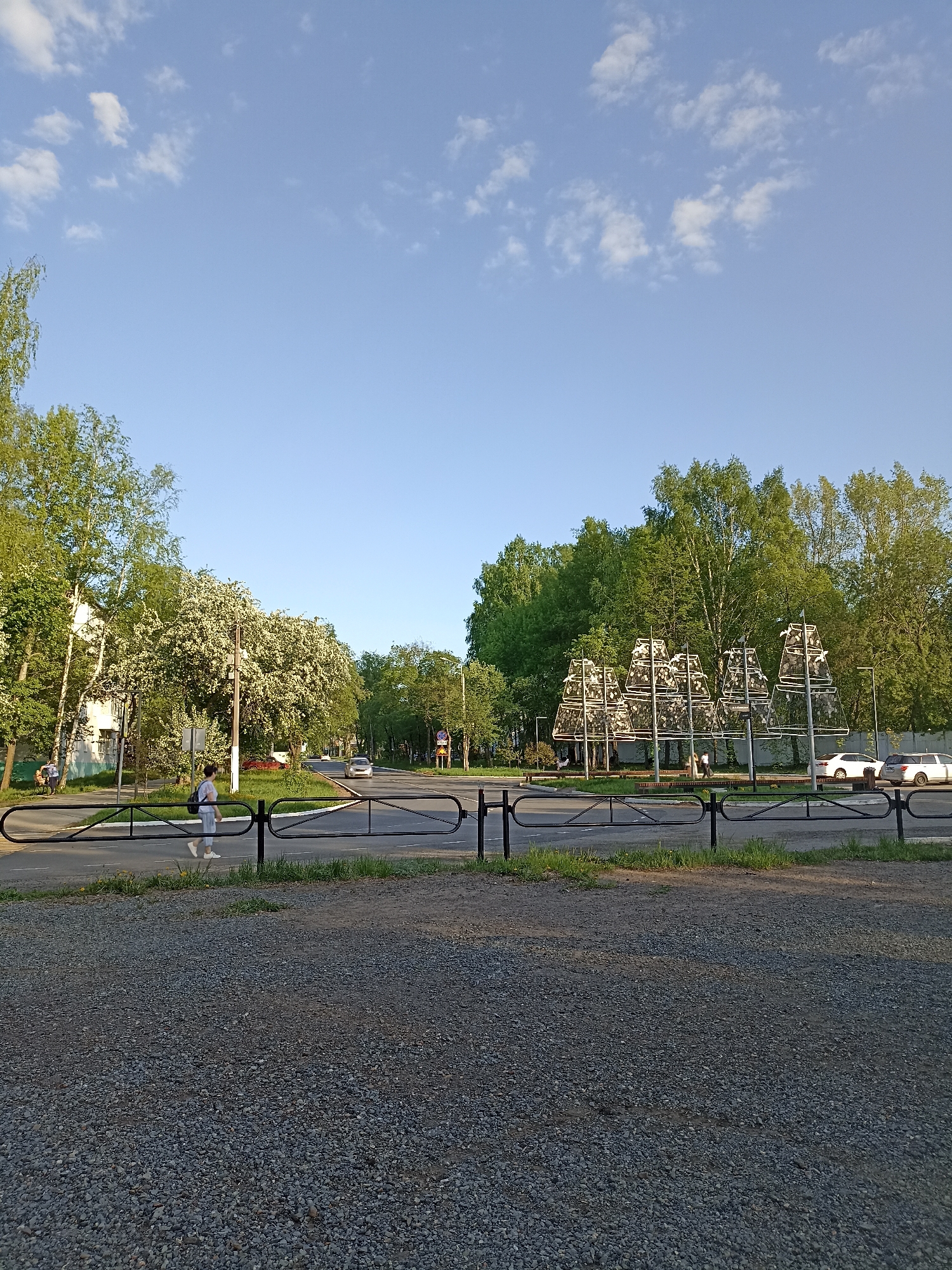The image depicts an outdoor scene in an urban environment. The sky is predominantly blue with scattered clouds. In the foreground, there is a gravel surface, followed by a road with a black metal fence running parallel to it. A person is walking on the crosswalk. Across the street, there is a park or green area with trees and bushes. A sculpture or art installation consisting of several pyramid-shaped structures is visible within the park. In the background, buildings and cars are visible, suggesting the location is a city.