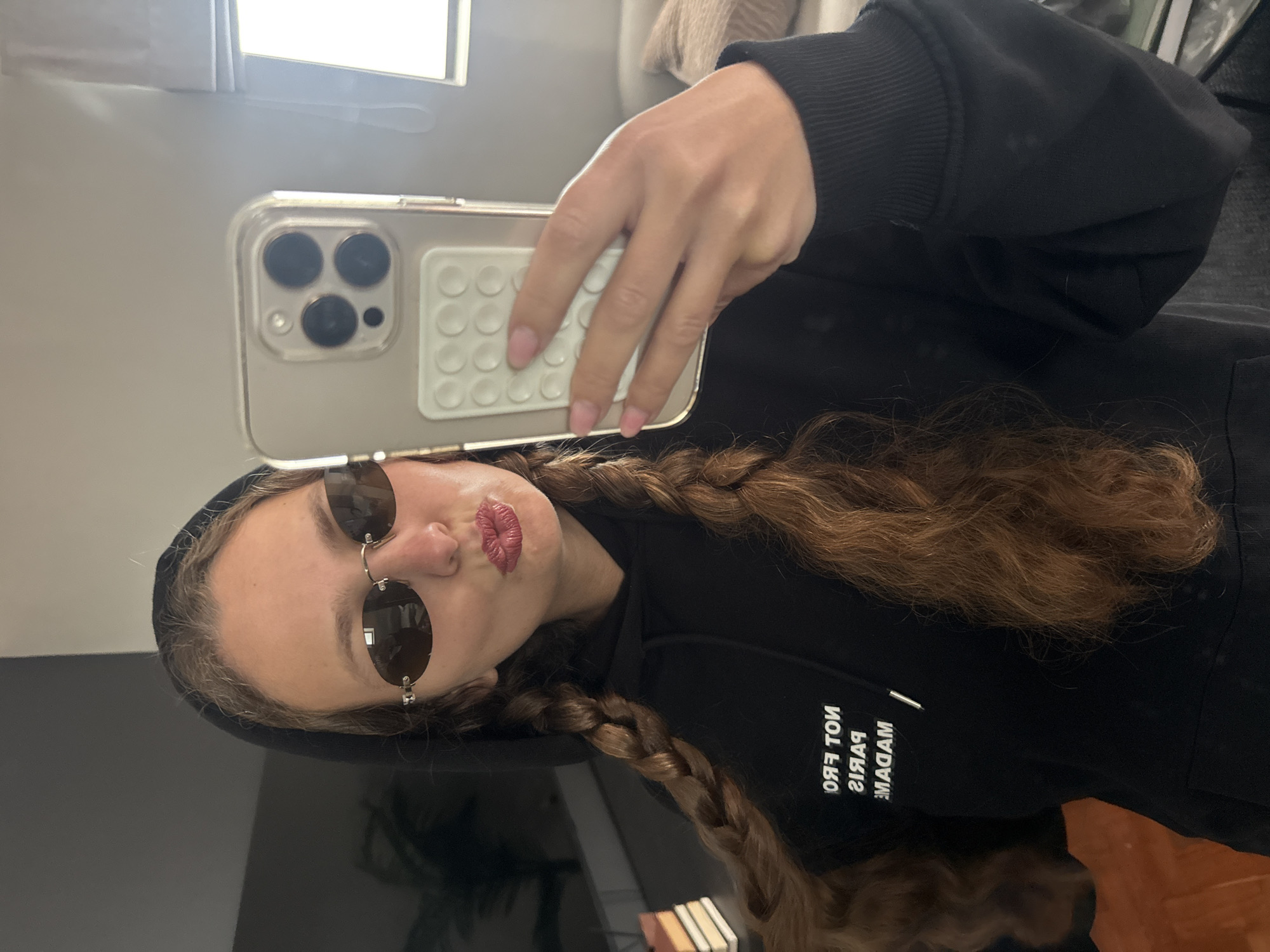 A selfie-style portrait captures a person with long, wavy brown hair styled into two braids, taken in Cape Town, South Africa. The individual is wearing a black hooded sweatshirt and dark, round sunglasses pushed down on their nose, revealing a puckered lip expression. They are holding a smartphone with a clear case and a white grip accessory (PopSocket-like) on the back. Light pink nail polish is visible on the fingers holding the device. The text on the black hoodie is seen in reverse, reading "NOT FROM PARIS MADAME" with a white vertical line element. The setting appears to be indoors, with a plain background wall and a bright light source at the top, possibly a window. A dark, reflective surface is visible in the lower part of the frame. The activity is clearly a self-portrait being taken in a casual setting.