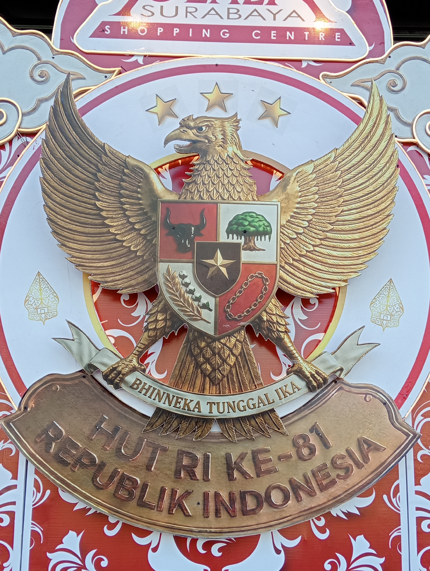 The image shows a close-up of a golden Garuda Pancasila emblem, the national emblem of Indonesia, displayed prominently on a red and white decorative background. The emblem features a mythical bird with outstretched wings, clutching a scroll in its talons. The scroll bears the national motto "Bhinneka Tunggal Ika" (Unity in Diversity). Below the Garuda, a bronze plaque reads "HUT RI KE-81 REPUBLIK INDONESIA," indicating a celebration of the 81st anniversary of the Republic of Indonesia. The backdrop includes stylized clouds and stars. The top of the image shows text that reads "SURABAYA SHOPPING CENTRE". This suggests the emblem is part of a celebratory display at the Surabaya Shopping Centre, likely for Indonesian Independence Day. There are no people visible in the image, and the focus is solely on the emblem and its immediate decorative elements.