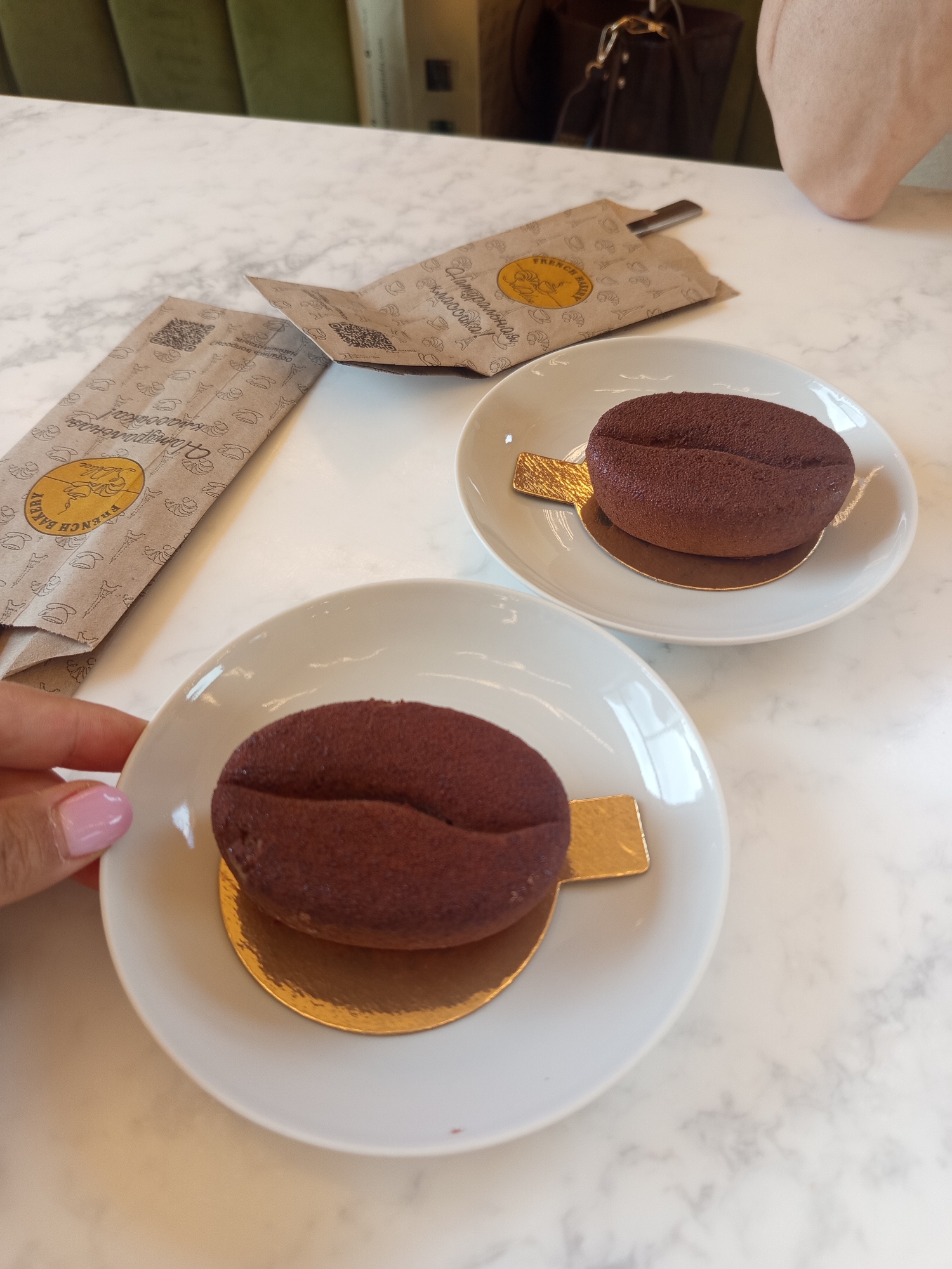 In the city of Misaylovo, Russia, two coffee bean-shaped chocolate cakes sit on white plates atop a marble-patterned table. Each cake is dusted with cocoa powder and rests on a golden base. Beside the plates are two brown paper bags with a French bakery logo and intricate patterns. A person's hand with pink nail polish is visible on the left, reaching for one of the plates. In the background, a blurred view of green upholstery and a dark handbag can be seen. The lighting suggests an indoor setting, possibly a cafe.