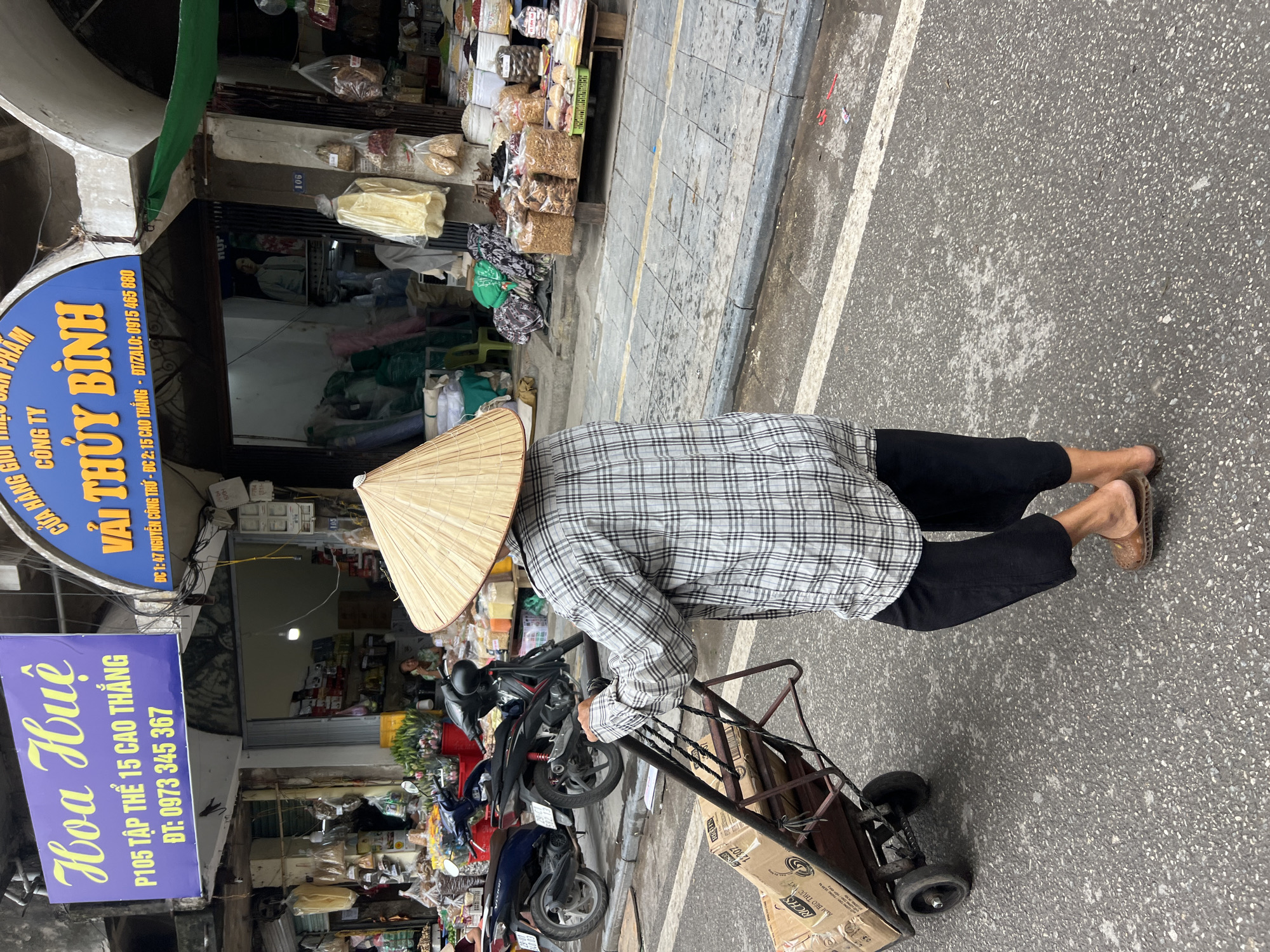 The image depicts a street scene in Hanoi, Vietnam, likely a market area. A person wearing a conical hat and a plaid shirt is pushing a two-wheeled cart laden with cardboard boxes. The cart has visible wheels and metal frame. In the background, several motorcycles are parked. Shops line the street, with various goods displayed, including bags of food items and fabrics. Signage in Vietnamese is visible on the buildings. The ground is paved with asphalt and stone tiles, with some areas appearing wet.