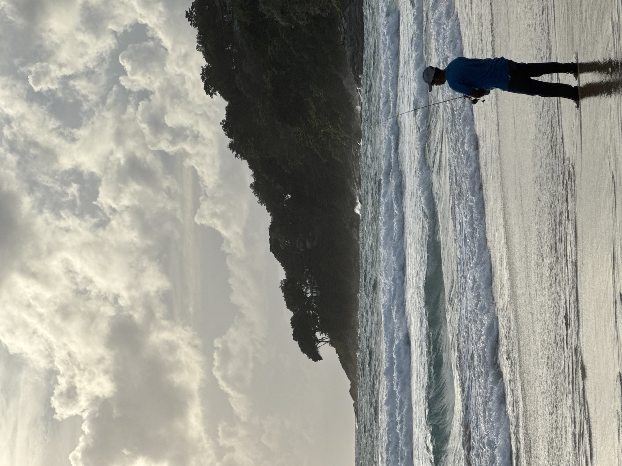 A person stands on a sandy beach in Karon, Thailand, fishing in the ocean. The person, silhouetted against the water, wears a blue t-shirt, dark pants, and a cap. They hold a fishing rod, casting into the waves. The ocean is rough with white-capped waves crashing onto the shore. In the background, a lush green, tree-covered hillside rises from the coast, partially obscured by dramatic, cloud-filled sky. The light suggests either early morning or late afternoon.