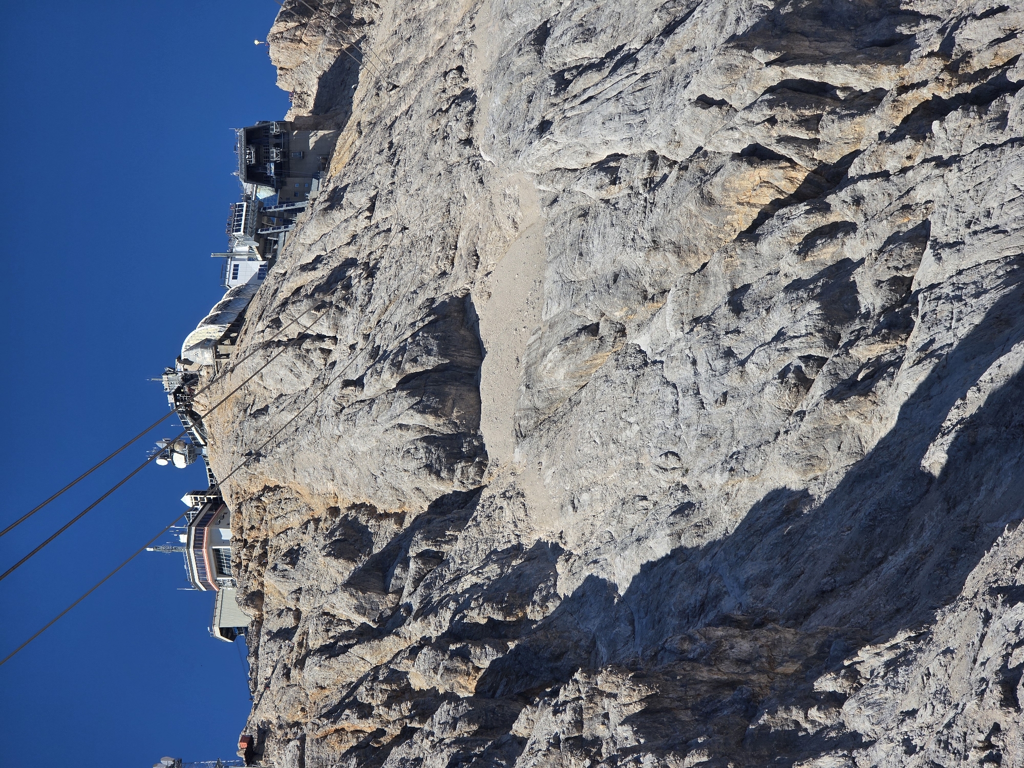 The image shows the Zugspitze summit station in Garmisch-Partenkirchen, Germany, as seen from below. The summit station is a complex of buildings perched atop a steep, rocky mountain face. Multiple cable car lines descend from the station, disappearing into the rugged terrain. The buildings themselves are modern, with large windows and metallic structures, and appear to house observation decks, restaurants, and other facilities. The mountain is characterized by its sharp, gray rock formations, with areas of scree and sparse vegetation. The sky is a clear, deep blue. There are no people visible in the image.