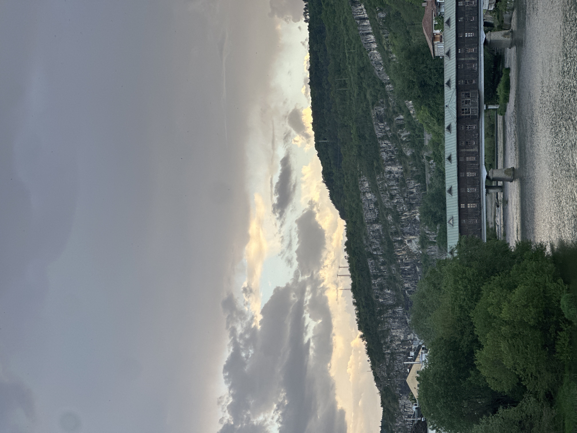 The image shows a landscape likely located in Lovech, Bulgaria, featuring a cloudy sky covering most of the view. To the right, there is a steep, rocky mountain covered in trees. A long, dark building with a green roof stands along the edge of the river next to the mountain. Part of the river can be seen on the right edge of the frame. In the foreground, there are more trees and some houses.