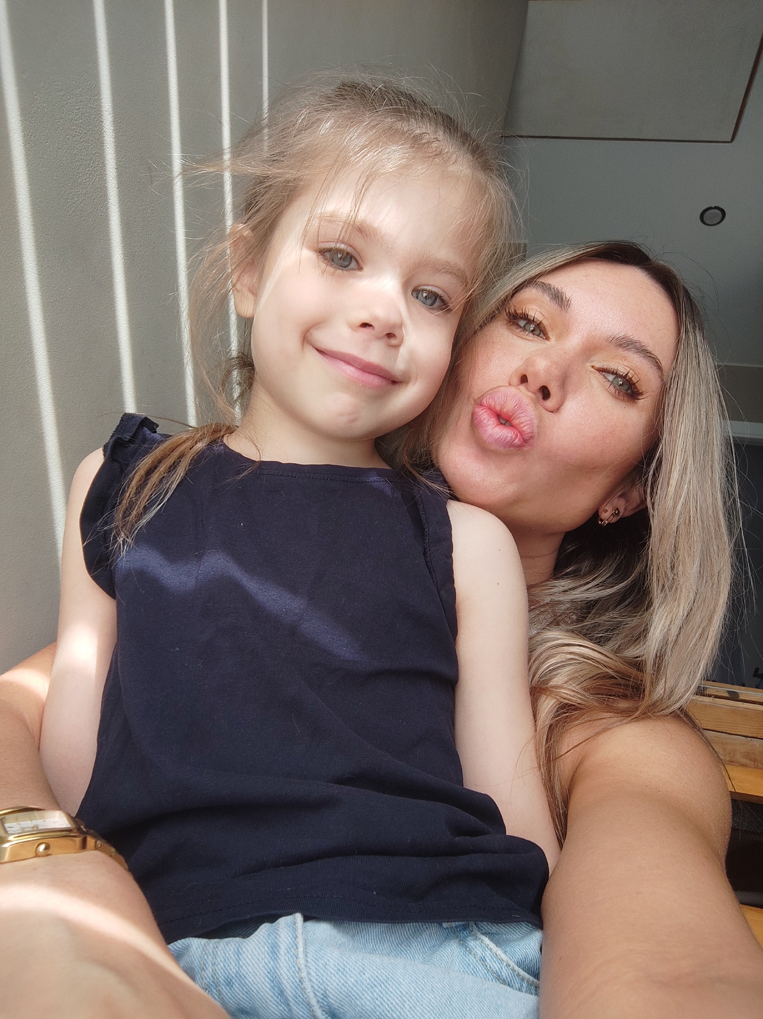 A woman with light blonde hair and a child are posing for a selfie. The child, who appears to be a girl, has light blonde hair, blue eyes, and is wearing a dark blue top. The woman is making a kissing face. They are indoors near a window with light streaming through, creating shadows on the wall behind them. The woman is wearing a gold watch and has pierced ears. Their clothes indicate they are wearing summer clothes. The location is not mentioned in the image and it is impossible to know this information.