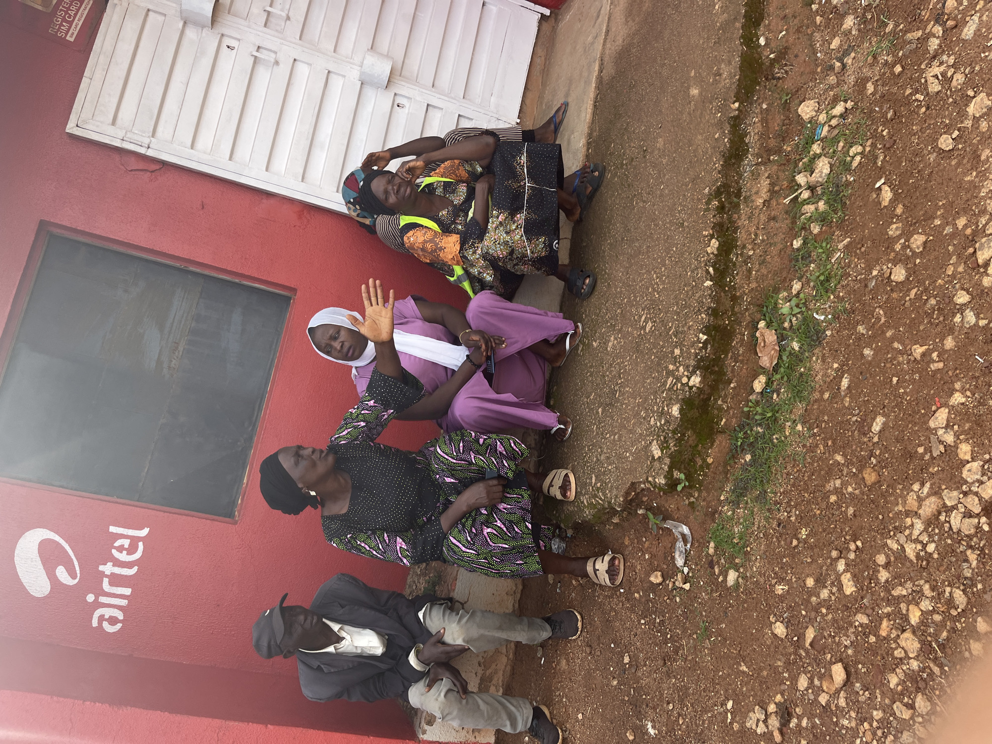 Four individuals, three women and one man, are seated on a concrete ledge in front of a red building with an Airtel logo. The man on the far left wears a grey jacket and a black cap. Next to him, a woman in a black patterned dress and headscarf gestures with her right hand. Another woman, in a purple dress and white headscarf, sits to her right. The fourth individual, a woman in a black and orange patterned dress and a yellow reflective vest, is seated on the far right, resting her head on her hand. The environment appears to be an outdoor urban setting with red-brown, rocky ground in the foreground. The building has a white, corrugated metal door and a window. A sign that reads "REGISTER SIM CARD" is visible above the door. The scene suggests a waiting area or a public space. The location is identified as Gura Top, Nigeria.