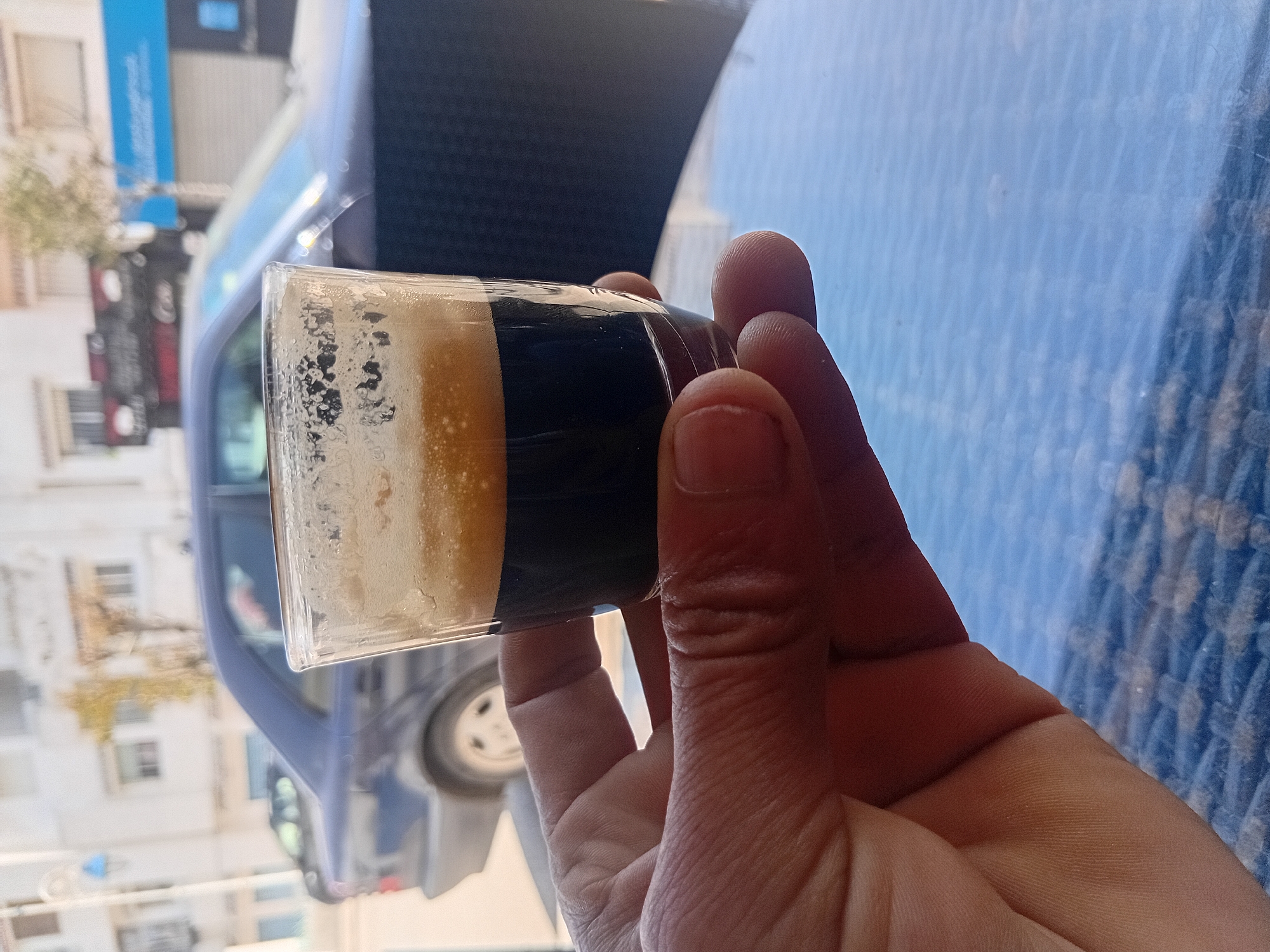 A hand holds a small glass of coffee, likely espresso, with a layer of crema on top. The scene is outdoors on a blue, patterned table, potentially at a café. A car is visible in the blurred background, parked on a street with buildings. The location is Martil ⵎⴰⵔْتⵉⵍ مرتيل, Morocco, suggested by the architectural style of the buildings and the overall ambiance.