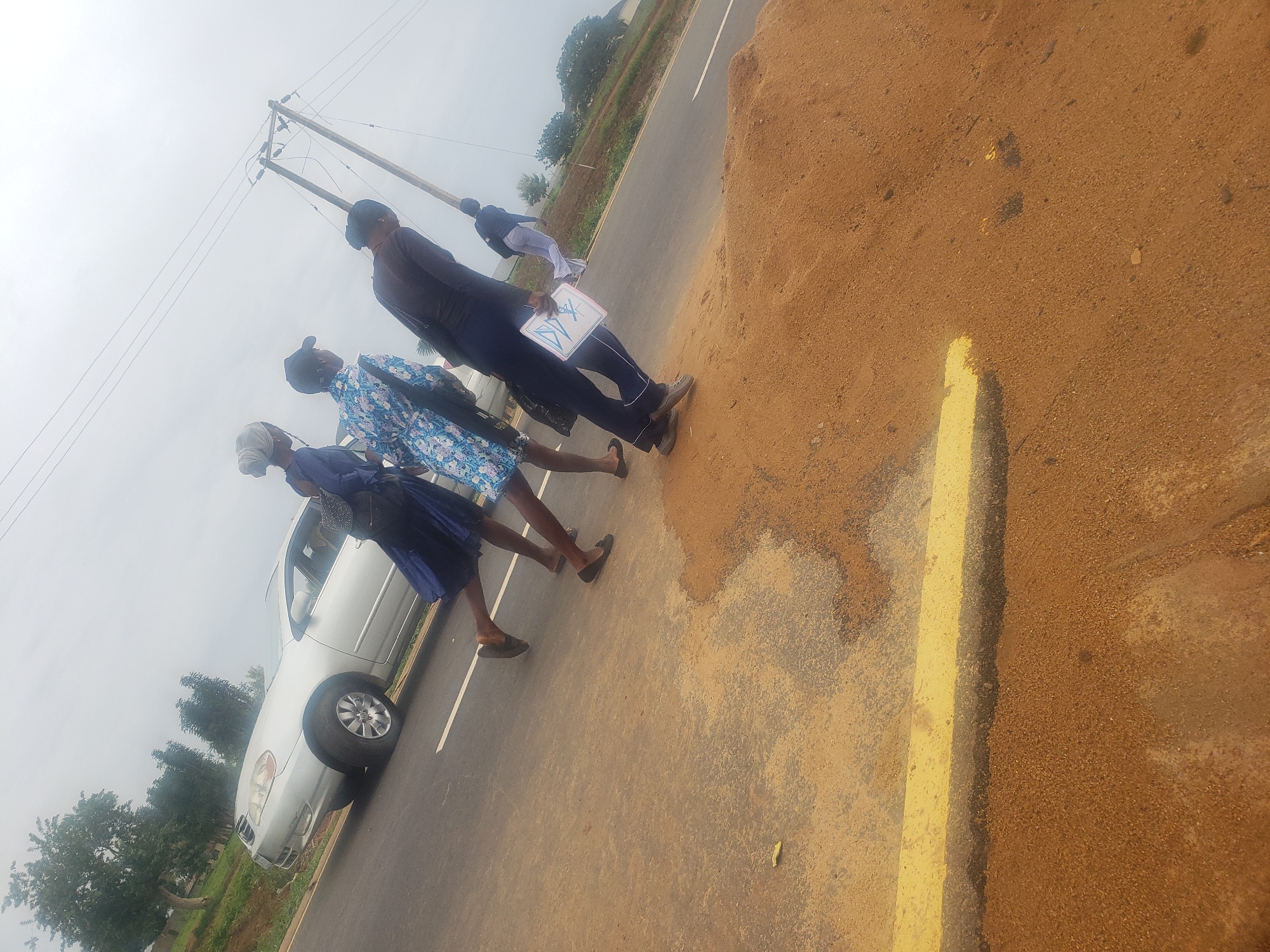 The image depicts a group of individuals on a paved road with a dirt embankment on the right side. In the background, a silver sedan is parked on the road. A utility pole with overhead wires stands to the right of the group. The sky is overcast. The people appear to be walking or standing near the road. The overall scene suggests an outdoor setting in Gumel, Nigeria.