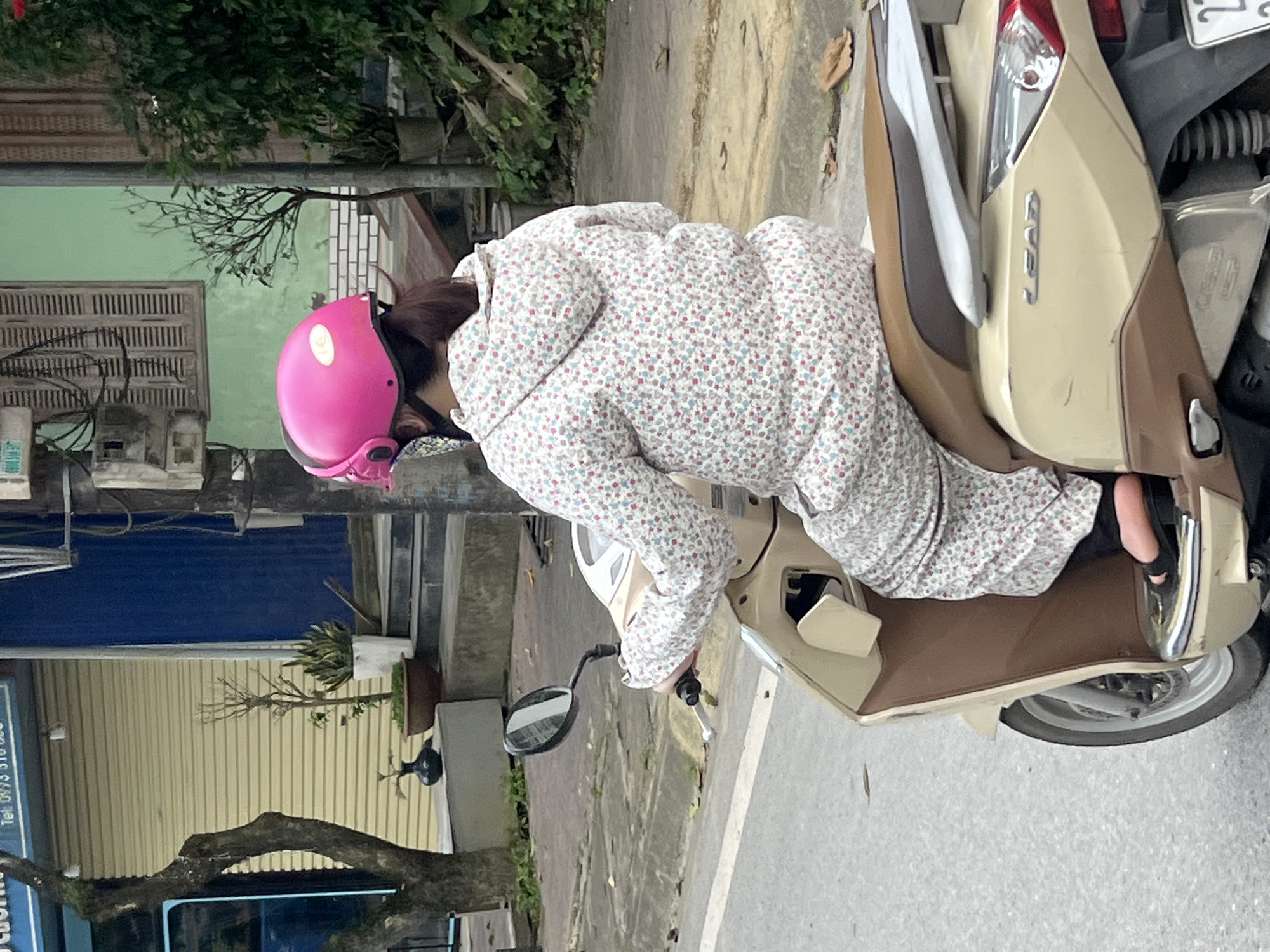A person wearing a pink helmet and a floral-patterned outfit is riding a beige scooter. The scooter is on a street in Hà Giang 2 Ward, Vietnam. The background shows a building with a yellow shuttered storefront, stairs, and lush greenery. The street appears to be paved, with a concrete curb and some fallen leaves. The overall scene depicts an everyday street view in a Vietnamese urban setting.