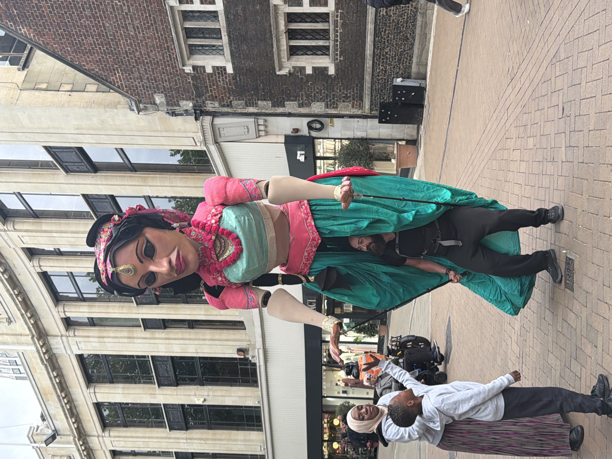 On a pedestrianized street in London, United Kingdom, a large puppet representing an Indian woman stands prominently. The puppet wears traditional Indian attire, including a pink and turquoise sari and ornate jewelry. A man dressed in black supports the puppet from underneath its skirt. Two people, a woman in a hijab and a young boy, interact with the puppet by reaching out to touch its hand. The street is paved with light-colored bricks, and several buildings line the street, with storefronts and windows visible. The architectural styles vary, with both modern and older brick buildings present.