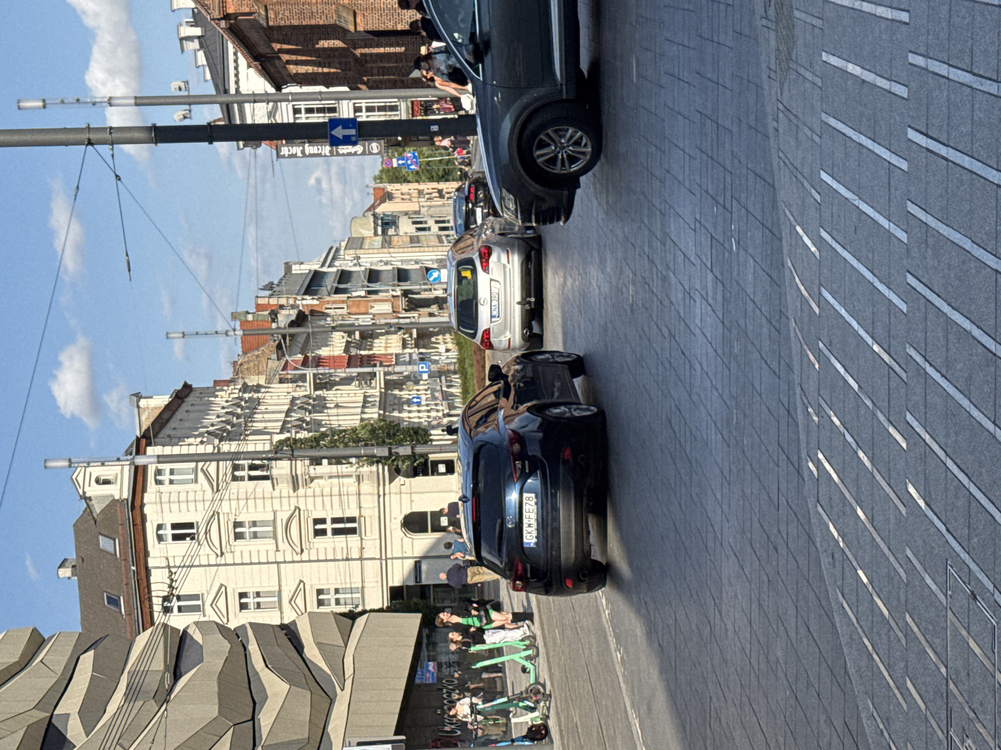 This is a medium-distance, eye-level, outdoor shot of a street scene in Poznan, Poland. The foreground is dominated by a dark blue Mazda 3 hatchback with the license plate "GKW-FE78", which is moving towards the viewer. To the right and slightly behind it, the front of a dark grey Audi Q7 SUV is visible, also moving forward. Further in the distance, a silver hatchback with the license plate "CNA 3907" is also on the road. The street is paved with grey interlocking tiles.
On the left side of the street, a modern, angular building with a grey facade is partially visible. Next to it are several parked electric scooters. A few people are standing near the scooters, and more pedestrians are visible further down the street.
The background features a row of traditional European-style buildings with light-colored facades, ornate windows, and red-tiled roofs. These buildings appear to be a mix of commercial and residential spaces. Overhead, a clear blue sky with a few scattered clouds is visible, along with power lines and street lamps. The overall lighting suggests it is daytime, with the sun casting shadows on the street.