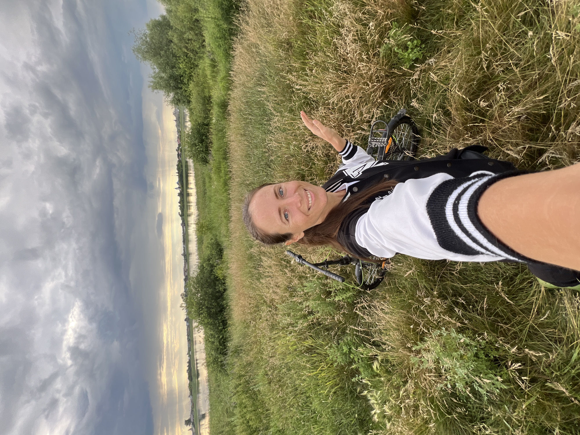 A medium shot shows a woman taking a selfie in a grassy field near the city of Pivkino, Russia. The woman, with her hair pulled back, is smiling at the camera and her right hand is extended, palm open, towards the right. She is wearing a black and white letterman jacket. Behind her, a bicycle is lying on its side in the tall grass. The background features a body of water, likely a river, and a distant treeline with some buildings visible. The sky above is filled with dramatic, dark clouds, with a sliver of sunlight breaking through on the horizon.