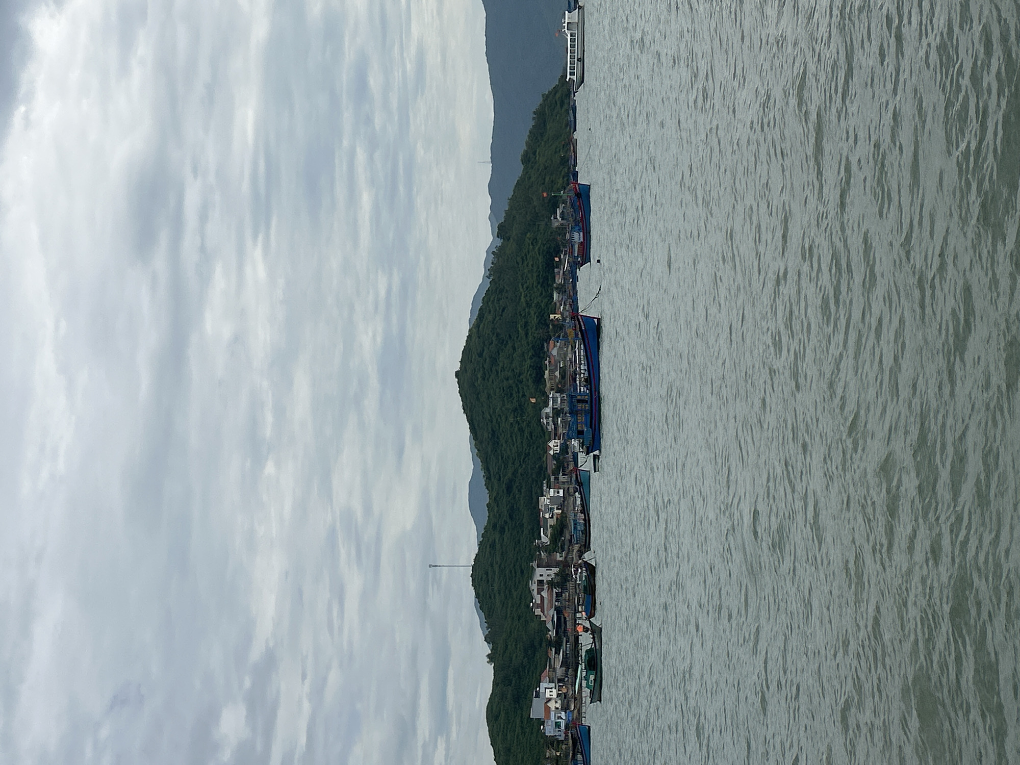 The image depicts a coastal scene in Ninh Ích, Vietnam, under a cloudy sky. The foreground is dominated by the choppy grey surface of the ocean. In the mid-ground, a fleet of fishing boats of varying sizes and colors are anchored or gently moving on the water. Several of these boats are painted in shades of blue, with some featuring red and yellow flags. A larger white passenger boat with multiple windows is visible on the far right.
The background features a densely vegetated, hilly coastline. Along the base of the hills, a town is nestled, characterized by a collection of buildings with white and red roofs, interspersed with darker structures. Some buildings appear to be multi-story residences or businesses. A prominent cell tower stands tall on one of the hills overlooking the town. The overall atmosphere is serene and maritime, indicative of a working fishing village or coastal town.