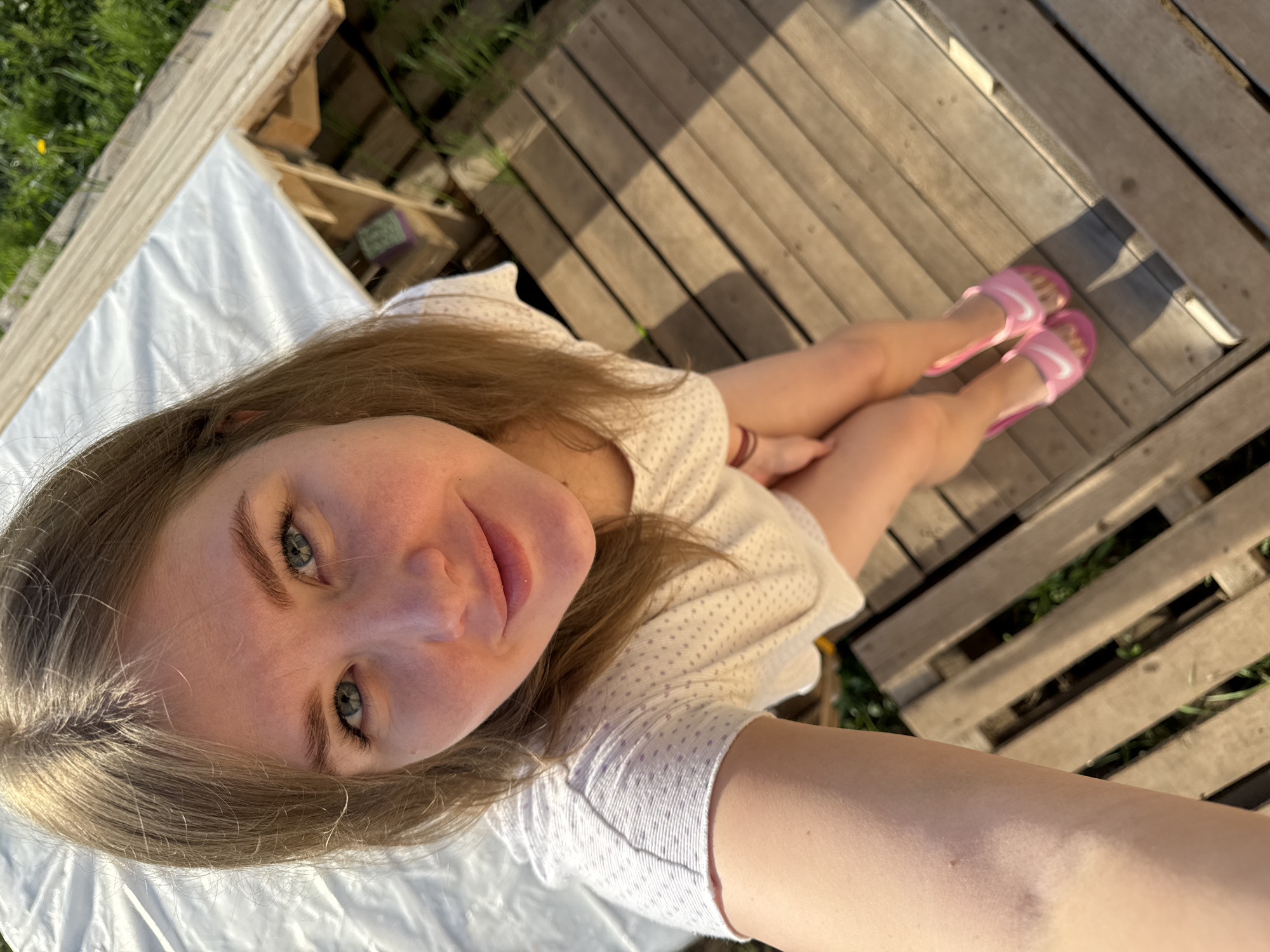 A young woman with blonde hair and blue eyes takes a selfie while sitting on a wooden pallet outside, presumably in Ферма, Russia. She wears a white t-shirt with small beige dots and pink Nike sandals. The sun casts shadows across the wooden pallet. A white sheet is visible to her left. The area appears to be outdoors with grass visible in the background.