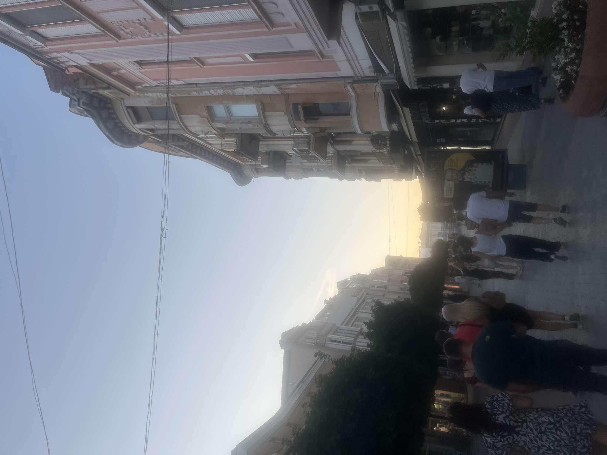 The image depicts a street scene in an urban environment, likely in the late afternoon or early evening, with a soft, warm light suggesting sunset. The perspective is from the street, looking down a pedestrian area lined with buildings on both sides.
On the right side of the street, a multi-story building with a distinct architectural style dominates the frame. It features ornate balconies, decorative moldings, and a mix of aged and painted facades, with sections in a pale pink color. The ground floor appears to house shops with awnings. A large poster featuring a portrait of a man in a military uniform is visible on the left side of the street, near the shops.
On the left side of the street, another building, partially obscured by trees, can be seen. The trees are dense and dark, casting shadows and creating a contrast with the brightly lit sky.
A number of people are walking along the street, mostly in the foreground and middle ground. They are dressed in casual summer clothing. Some are walking alone, while others are in groups. Their faces are not clearly discernible due to the image quality and distance. The pavement is visible, and a large planter with flowers is positioned in the lower right corner. Overhead, power lines are strung across the sky. The sky itself is a pale blue, transitioning to a lighter, hazy hue towards the horizon where the sun is likely setting.