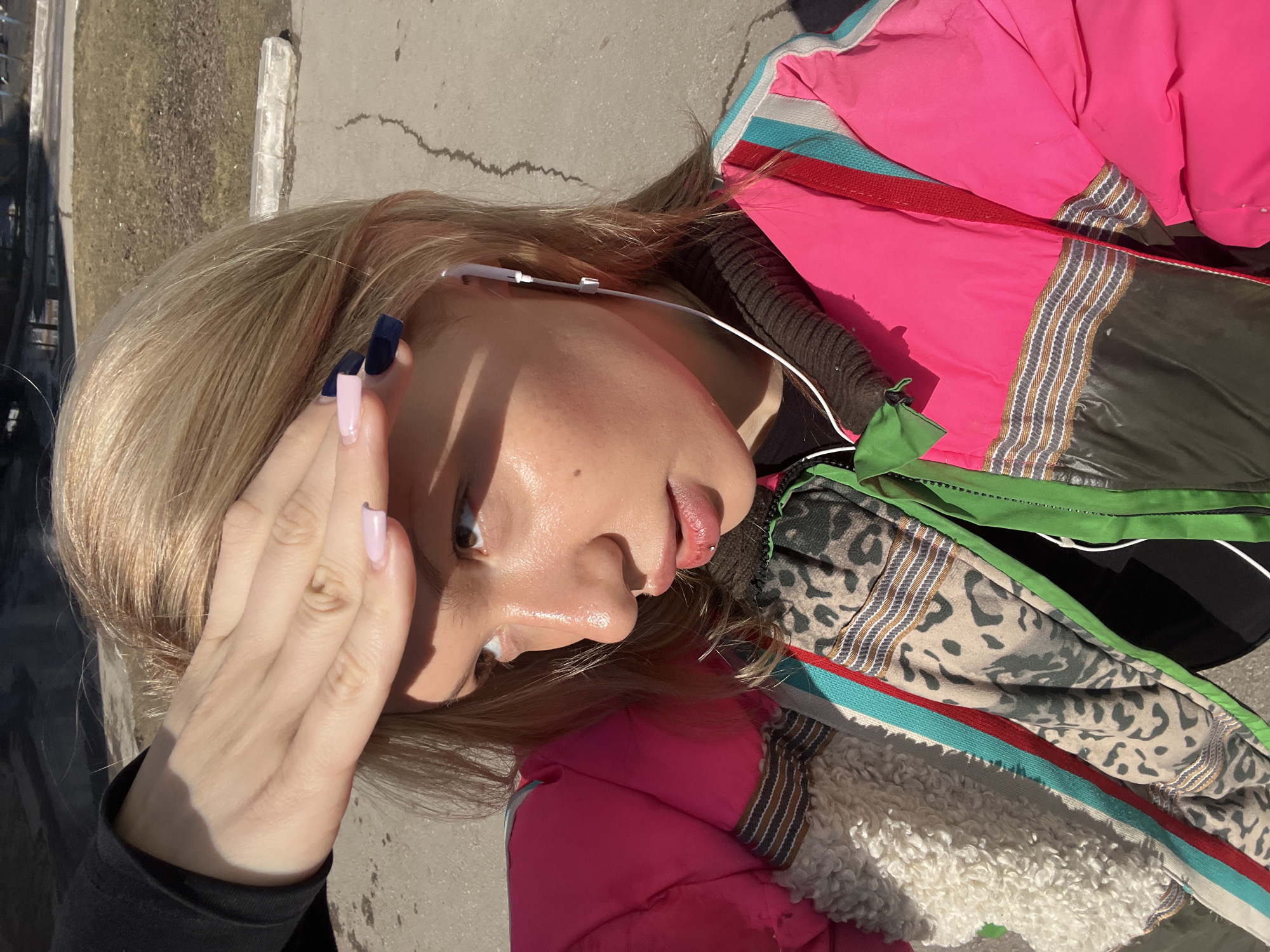 The image depicts a young woman with fair skin and light blonde hair, likely taken outdoors in Chaikovsky, Russia. She is wearing a multicolored jacket with elements of pink, green, brown, black, and white. She has earphones in her ear. Her nails are painted in a color-block style with blue and pink shades. She is positioned near a grey concrete surface, possibly a ledge or sidewalk. The lighting is bright, suggesting daylight. She has her hand near her face and looks towards the right.