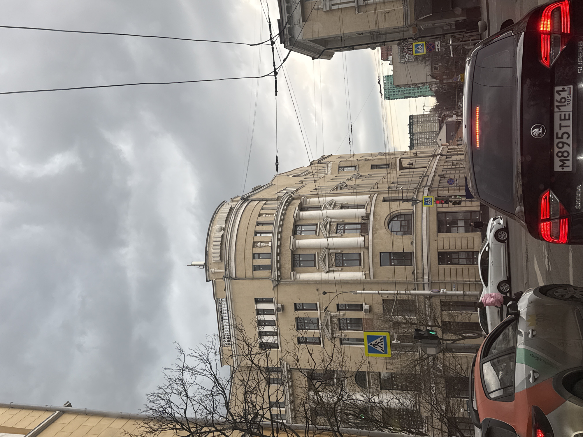 The image shows a street scene in Rostov-on-Don, Russia. The architectural style of the tan building suggests a historic or possibly Soviet-era structure. A black Skoda car is immediately visible. Pedestrian crosswalk signs are present. Power lines are overhead. Traffic is present. Bare tree branches suggest it's late fall or early spring. The sky is overcast. A few pedestrians can be seen. The license plate on the Skoda is M895TE 161 RUS.