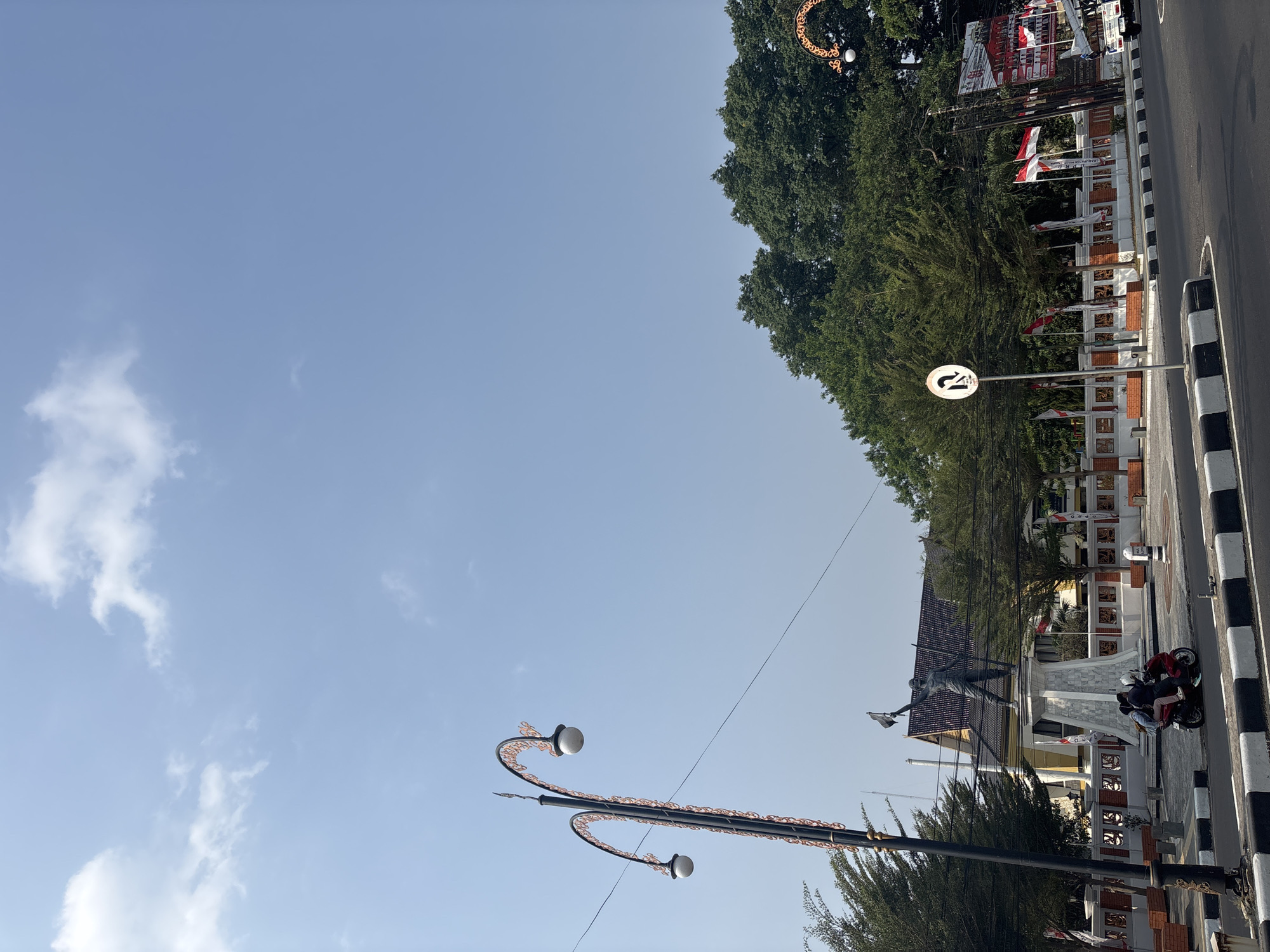 A statue of a man holding a flag stands on a pedestal in a public square, possibly in Majalengka, Indonesia, given the prevalence of Indonesian flags. The statue is surrounded by trees, fences, and streetlights. Two people ride a red scooter in the foreground, and a white truck is visible in the background. The sky is clear and blue with a few scattered clouds.