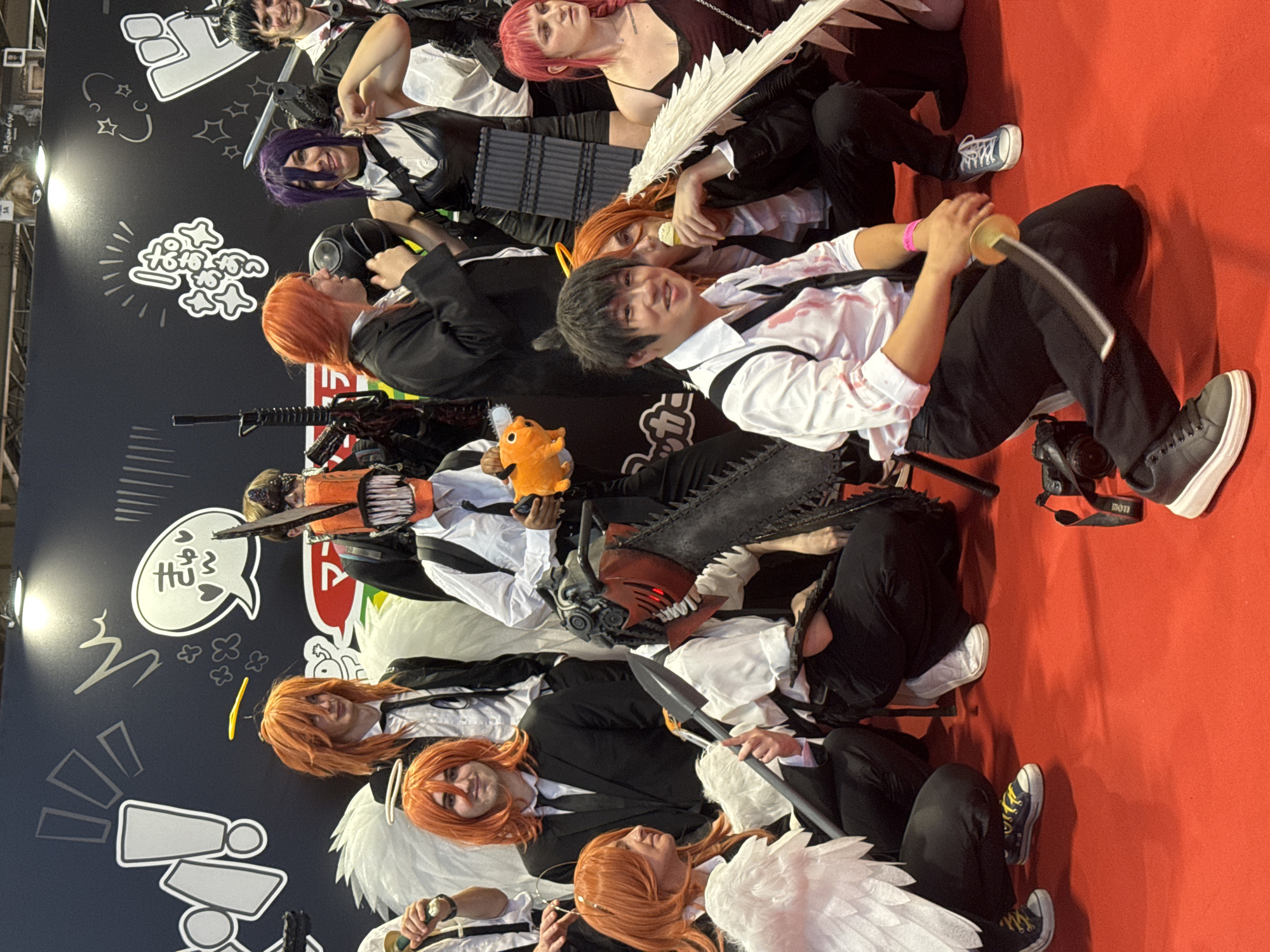 At the Paris Nord Villepinte Exhibition Centre in Villepinte, France, a group of cosplayers, likely attending a convention, are posing for a photograph on a bright red carpet. The backdrop features a black wall with white graphic designs, possibly related to a manga or anime series. The individuals are dressed in costumes, many of whom have orange hair and appear to be portraying characters from "Chainsaw Man," including Denji (with a chainsaw head), Aki Hayakawa, and possibly Power. Some are wearing formal attire like suits and ties, with blood splatters on some of their white shirts. One person is holding a plush toy of Pochita, the chainsaw dog. Another individual is brandishing a prop sword. Several people are wearing white feathered wings. The lighting suggests an indoor convention environment.