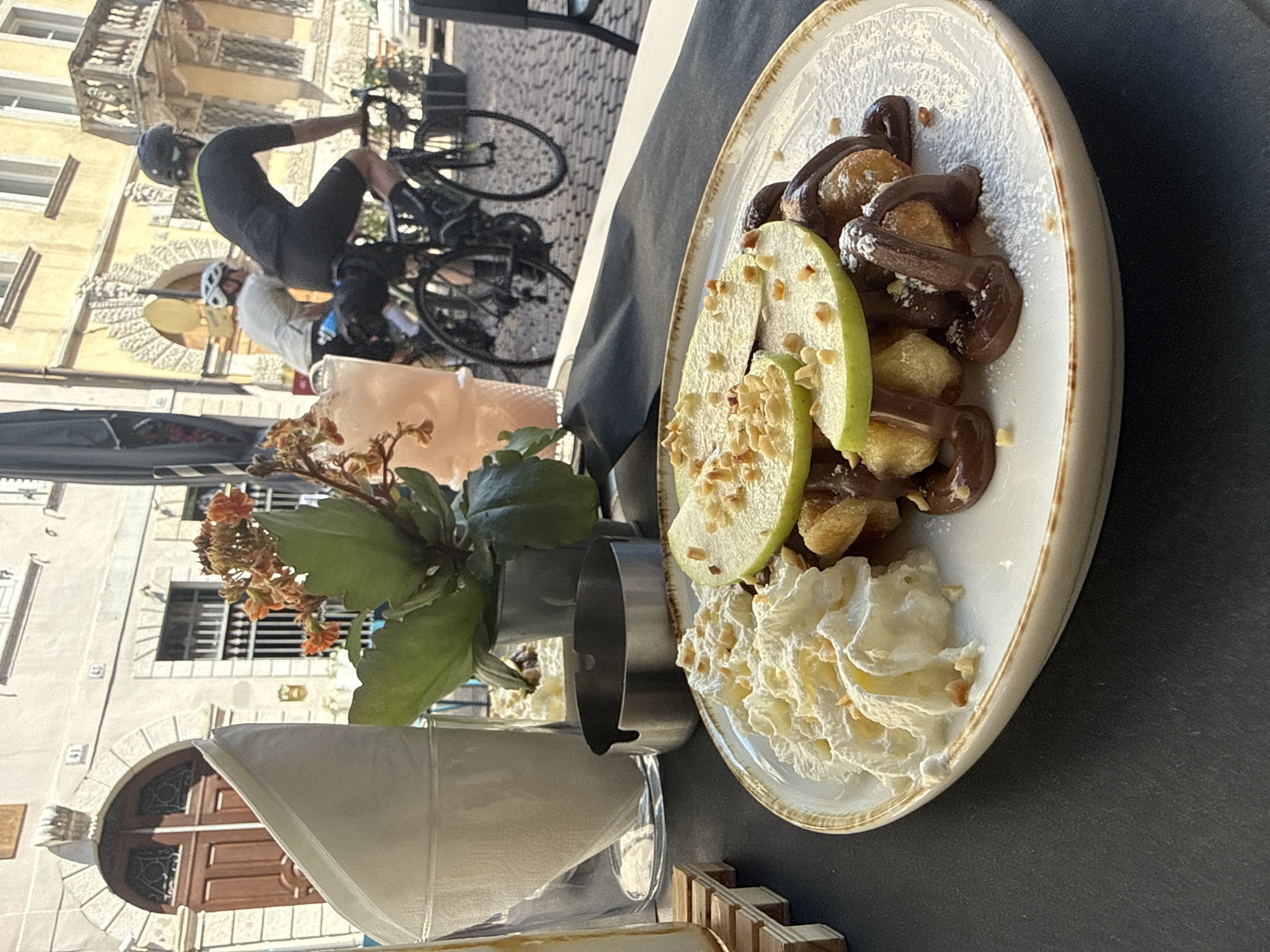 The image captures a close-up view of a dessert on a white plate with a rustic rim, placed on a dark surface. The dessert consists of small, golden-brown pieces, possibly mini waffles or pastries, generously drizzled with melted chocolate and dusted with powdered sugar. Slices of green apple, topped with chopped nuts, are artfully arranged on the dessert. To the left of the plate, a swirl of whipped cream, also sprinkled with nuts, is presented. A metallic ashtray sits behind the dessert, and a glass containing a pale pink beverage and a folded white napkin are visible further back. In the background, two individuals on bicycles are partially visible. One cyclist, wearing a black cycling outfit and helmet, is in the foreground, while another, in a lighter-colored top and helmet, is behind them. They are riding on a cobblestone street. The architecture in the background suggests an urban setting with stone buildings featuring arched doorways and windows. The overall scene appears to be an outdoor cafe in Trento, Italy, given the architectural style and the presence of cyclists, which is common in Italian cities. The lighting indicates it is daytime, with sunlight illuminating the scene.