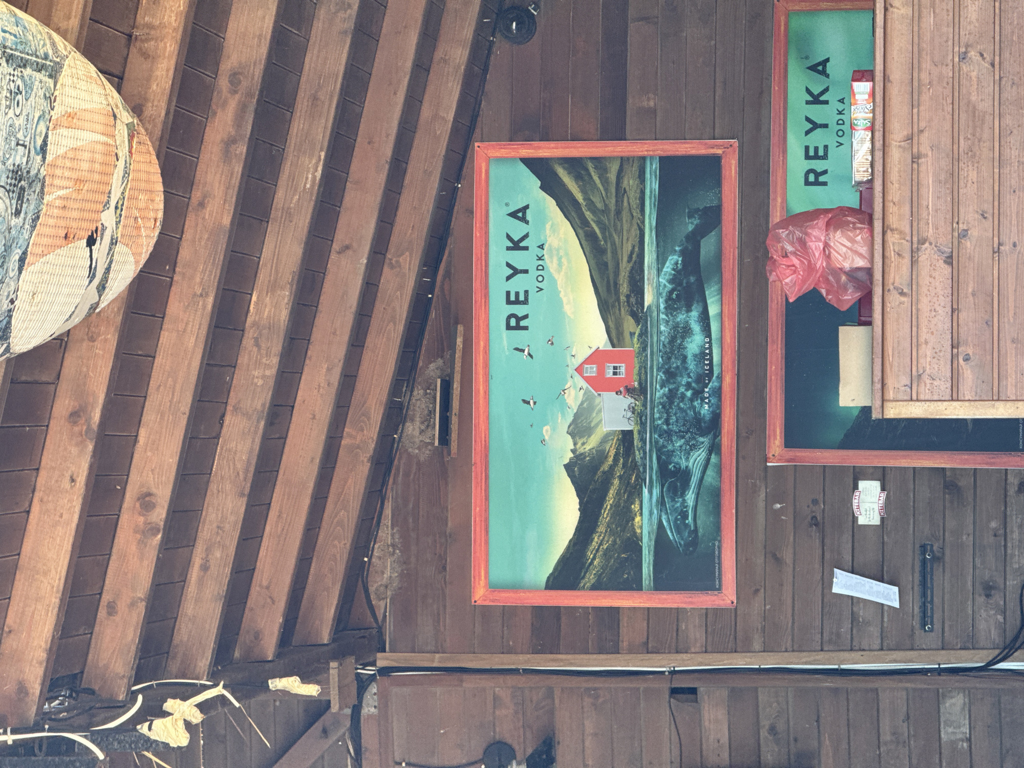The scene is set within a wooden-paneled structure, likely an outdoor covered area or interior establishment, in Sozopol, Bulgaria. The walls are constructed from horizontal dark-stained wooden planks, while the ceiling features parallel, wider wooden beams supporting narrower wooden planks.
A prominent, large rectangular sign advertising "REYKA VODKA" is mounted centrally on the wooden wall. This sign is framed in a reddish-orange border. Its artwork depicts a stylized Icelandic landscape: a light blue sky with several flying birds (gulls), green mountainous terrain, a small red house on an islet, and a person near a bicycle next to a white structure. Below the waterline, the lower portion of a large whale is visible. The text "MADE OF ICELAND" and partially obscured text "KONSÚMÍRAI OFFLINE" are visible at the bottom of the sign.
Directly below the large sign, a smaller, similarly framed rectangular sign also displays "REYKA VODKA" against a green background. A wooden shelf or counter extends horizontally in front of this lower sign. On this shelf, from left to right, are a light-colored rectangular object (possibly a box), a crumpled red plastic bag, and a white and red rectangular box (possibly containing snacks or crackers).
To the left of the lower sign, a white piece of paper is taped diagonally to the wooden wall. Below it, a short horizontal metal bar with two rings is affixed to the wall. Above this bar, two small stickers are present, one clearly legible as "KITEBAR". Visible on the vertical wooden planking to the far left is a black circular fixture, possibly a speaker or a light. Another identical circular fixture is mounted on the wall to the right of the upper Reyka sign.
The ceiling displays visible electrical wiring running along the wooden beams. In the upper right quadrant of the image, a large, decorative, multi-colored woven object, resembling a basket or lampshade, hangs from the ceiling structure. No actual people are present within the immediate visible area, though a small figure is depicted within the artwork of the upper Reyka sign.