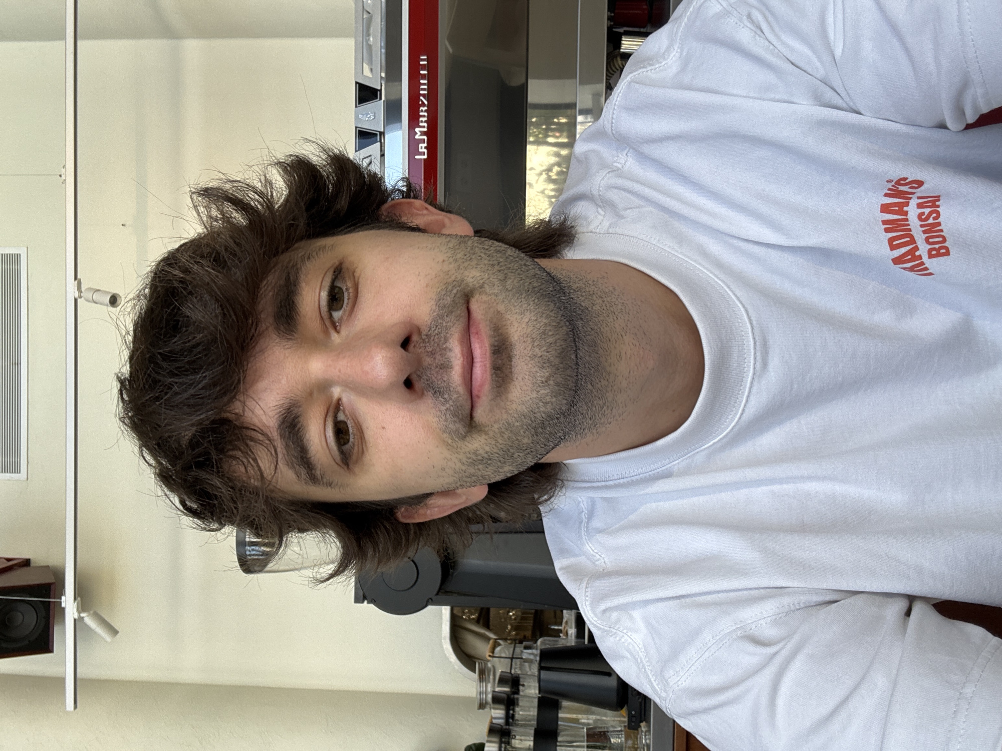 This is a selfie of a young man with dark, wavy hair and a light beard. He is wearing a white t-shirt with a red logo that reads "MADMAN'S BONSAI". He is looking directly at the camera with a neutral expression. Behind him, a red and silver La Marzocco espresso machine is visible, along with some shelving and equipment, suggesting he is in a cafe or coffee shop. The lighting appears to be artificial, possibly indoors during the day. There are also some other objects in the background, including a speaker and some shelving units. The image is oriented vertically, with the subject's head at the bottom and his feet at the top.