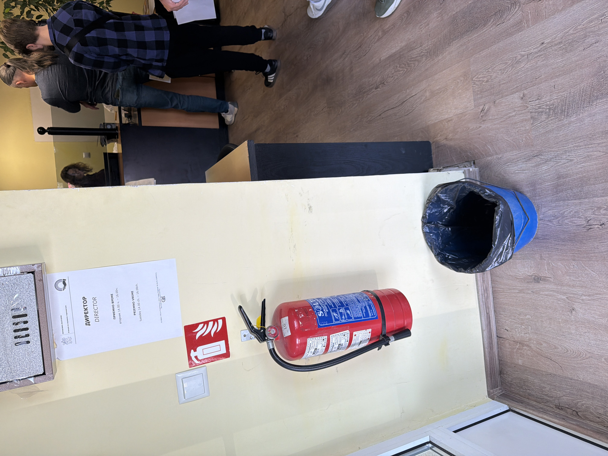 The image shows an interior scene with several key elements. A fire extinguisher is mounted on a beige wall, next to a red fire safety sign and a light switch. Above the fire extinguisher and sign, a document is partially inserted into a silver metal box or frame, with text in Bulgarian and English, "ДИРЕКТОР DIRECTOR," and mentioning "PRIEMNO VREME" and "tuesday 14.00 ch. - 16.00ch."
To the right of the fire extinguisher, a blue trash can with a black trash bag sits on the wooden floor. In the background, there are several people, one wearing a blue and black plaid shirt. The floor is made of wooden planks.
Without further information such as identifying signs outside the building, determining the exact location in Sofia, Bulgaria, is not possible.