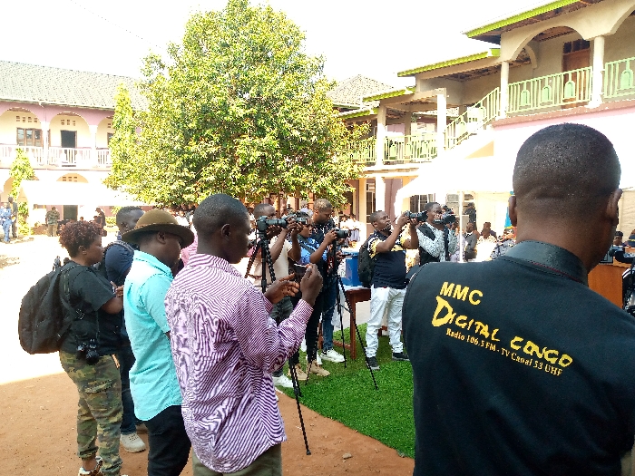 A group of about 15 people stand in the courtyard of a building in Beni, Democratic Republic of Congo. They are all focused on taking pictures with their cameras. The courtyard is covered in dirt and surrounded by buildings and trees. One person is wearing a black shirt with yellow writing that says “MMC Digital Congo, Radio 106.5 FM, TV Canal 53 UHF." The building behind the group is a two-story pink building with a white roof. Behind the building, there is a tall tree with green leaves. The sky is blue and mostly cloudless.