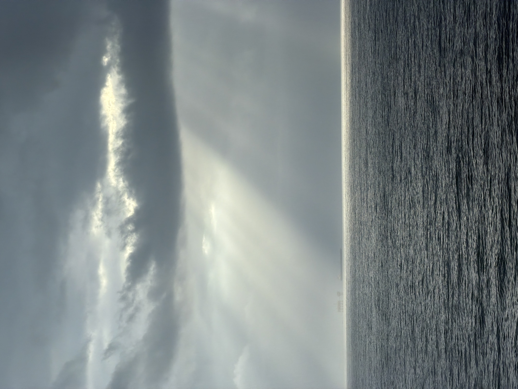 The image displays a vast seascape under a dramatic, cloudy sky, originally oriented vertically but best interpreted horizontally. A clear horizon line divides the expansive sky from the large body of water.
The sky is filled with heavy, dark clouds, but dynamic light breaks through in several places. Prominent crepuscular rays pierce the clouds, casting bright, ethereal shafts of light downwards and illuminating parts of the cloud formations. Some clouds are intensely bright where the light hits, while others remain dark and heavy, suggesting a powerful, unseen light source, likely the sun.
The surface of the water, which appears to be an ocean or a very large sea, is dark and highly textured. Countless small waves and ripples create a pattern of intricate lines across its surface, conveying a sense of immense scale and depth. The water's coloration is a deep, dark gray-blue.
On the distant horizon line, faint and indistinct structures are visible. These small, dark silhouettes could be ships, offshore platforms, or distant land formations/buildings, barely discernible through atmospheric haze or distance.
The scene captures a powerful and atmospheric moment, possibly during sunrise, sunset, or a dramatic break in overcast weather. The overall mood is one of solemn grandeur and vastness. There are no people visible, nor any discernible text.