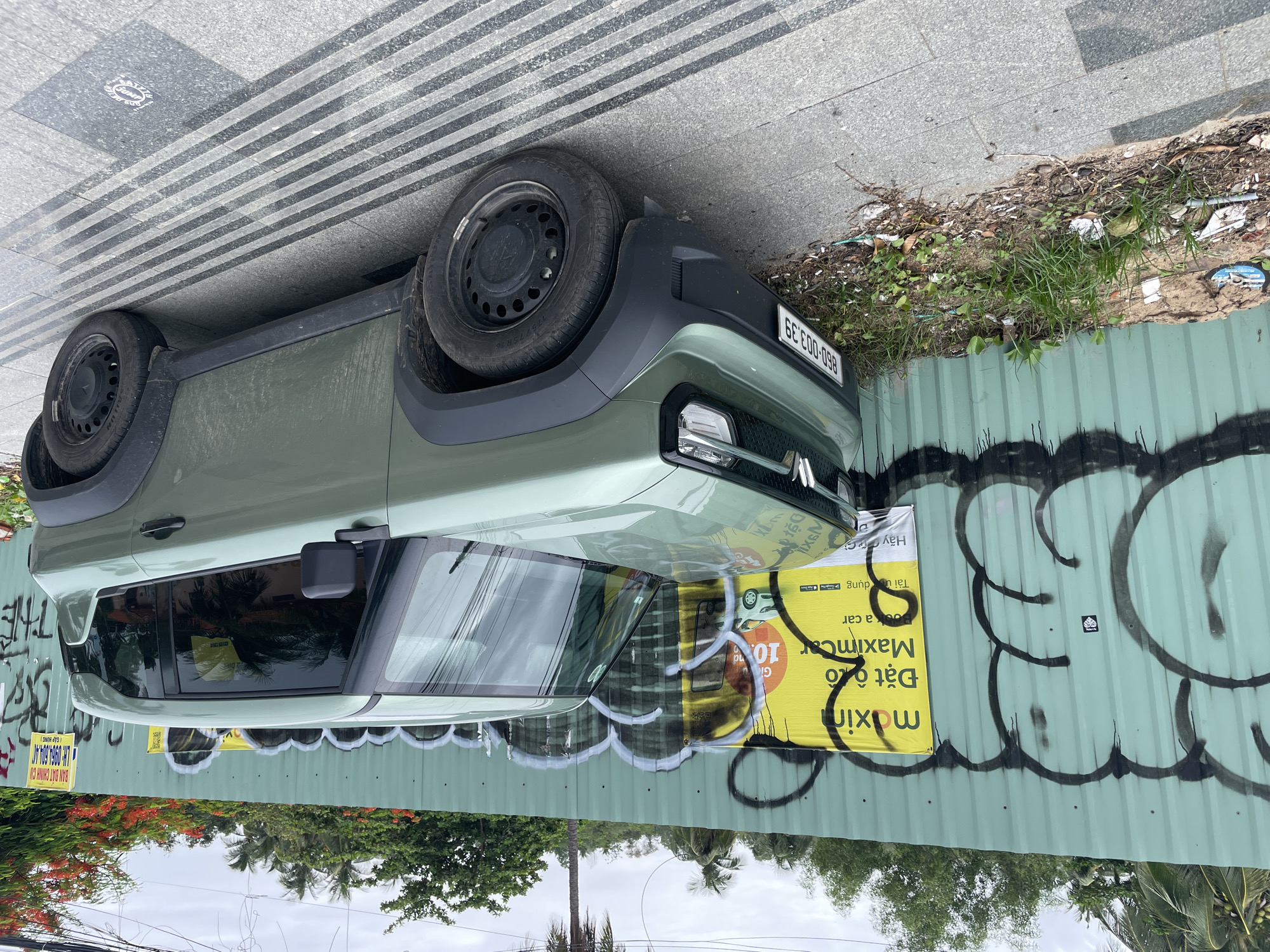 The image shows a light green Citroen Ami parked next to a corrugated metal fence in what appears to be a street setting. The fence is adorned with black graffiti and a yellow advertisement poster for "maximcar". The advertisement includes Vietnamese text, the word "Maximcar" in a yellow font, and the English words "book a car" and "Download app". A license plate is visible on the car. The ground consists of both pavement and a patch of grass and weeds, with some scattered trash. Trees are visible in the background, suggesting a residential or commercial area. The image is likely taken in Phu Thuy Ward, Vietnam, based on the OCR text "BAN DAT CHINH CU (GAPHONG)" which indicates a real estate service located in that ward.
