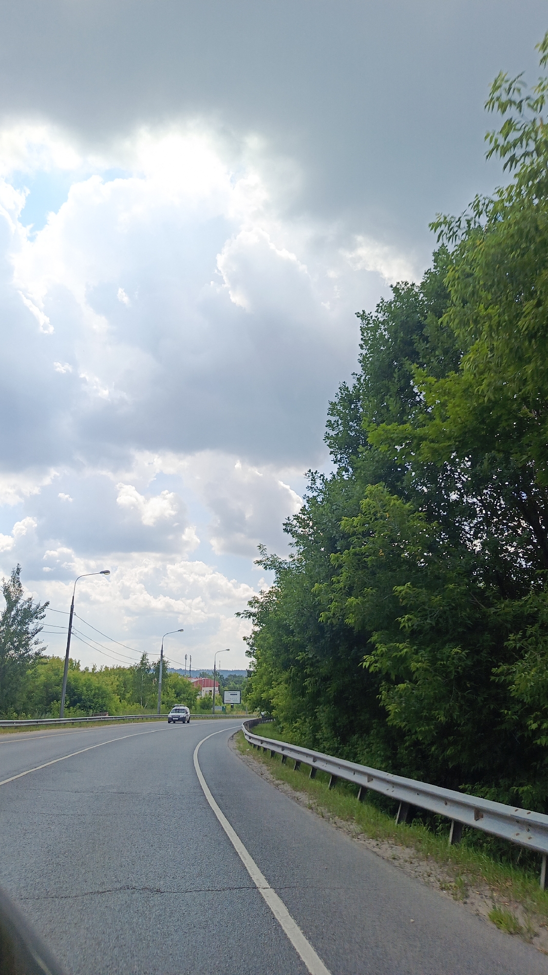 The image shows a view from a vehicle traveling on a paved road. The road curves to the right, with a metal guardrail on the right side. On the left side of the road, there are trees and streetlights. In the distance, there are more trees, some buildings, and a sign. A white car is visible driving on the road in the distance.
The sky is filled with clouds, with patches of blue sky visible. The sun appears to be shining through the clouds, creating bright areas. This suggests it is daytime, likely late morning or afternoon. The lush green trees indicate that it is likely summer.
The overall scene is one of a suburban or rural landscape, with a road leading through a wooded area. The mood is calm and serene. There is no visible text in the image.