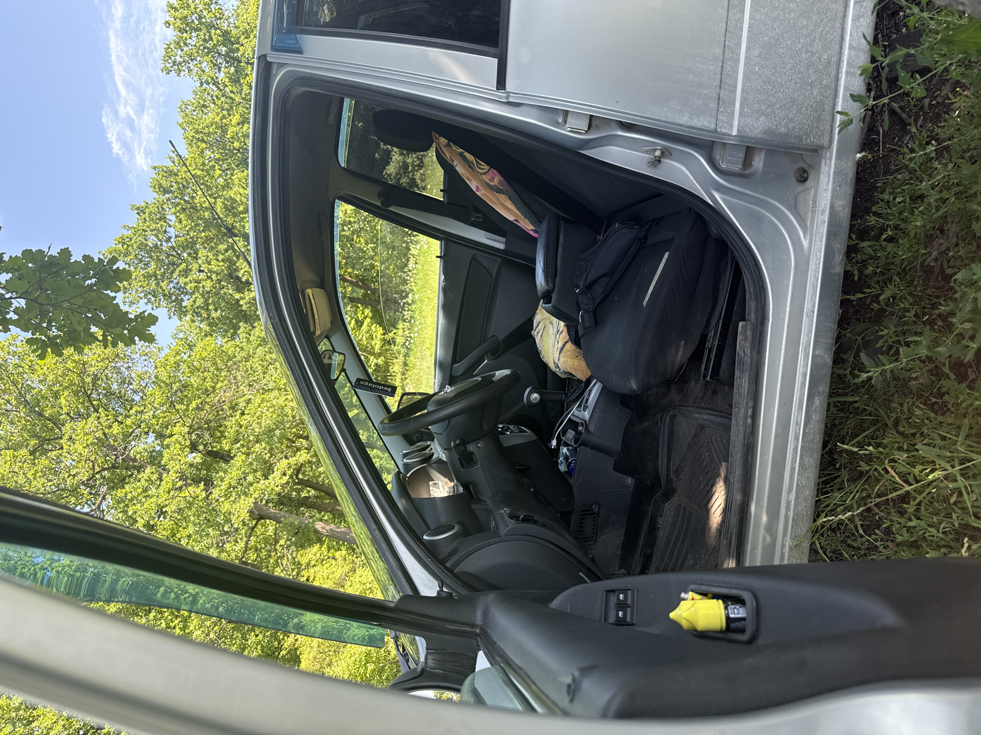 The image shows the interior of a silver car, parked outdoors. The driver's side door is open, revealing the front seats and dashboard. The car is surrounded by lush green trees and vegetation, with a blue sky and white clouds visible in the background. The interior of the car appears cluttered, with various items on the seats and floor. A yellow object, possibly a spray bottle, is visible on the inside of the driver's door. The overall scene suggests the car is parked in a natural setting, possibly a park or a wooded area. There are no people visible in the image. The image does not provide any specific details or clues that would help identify its location in the city of Samara, Russia.