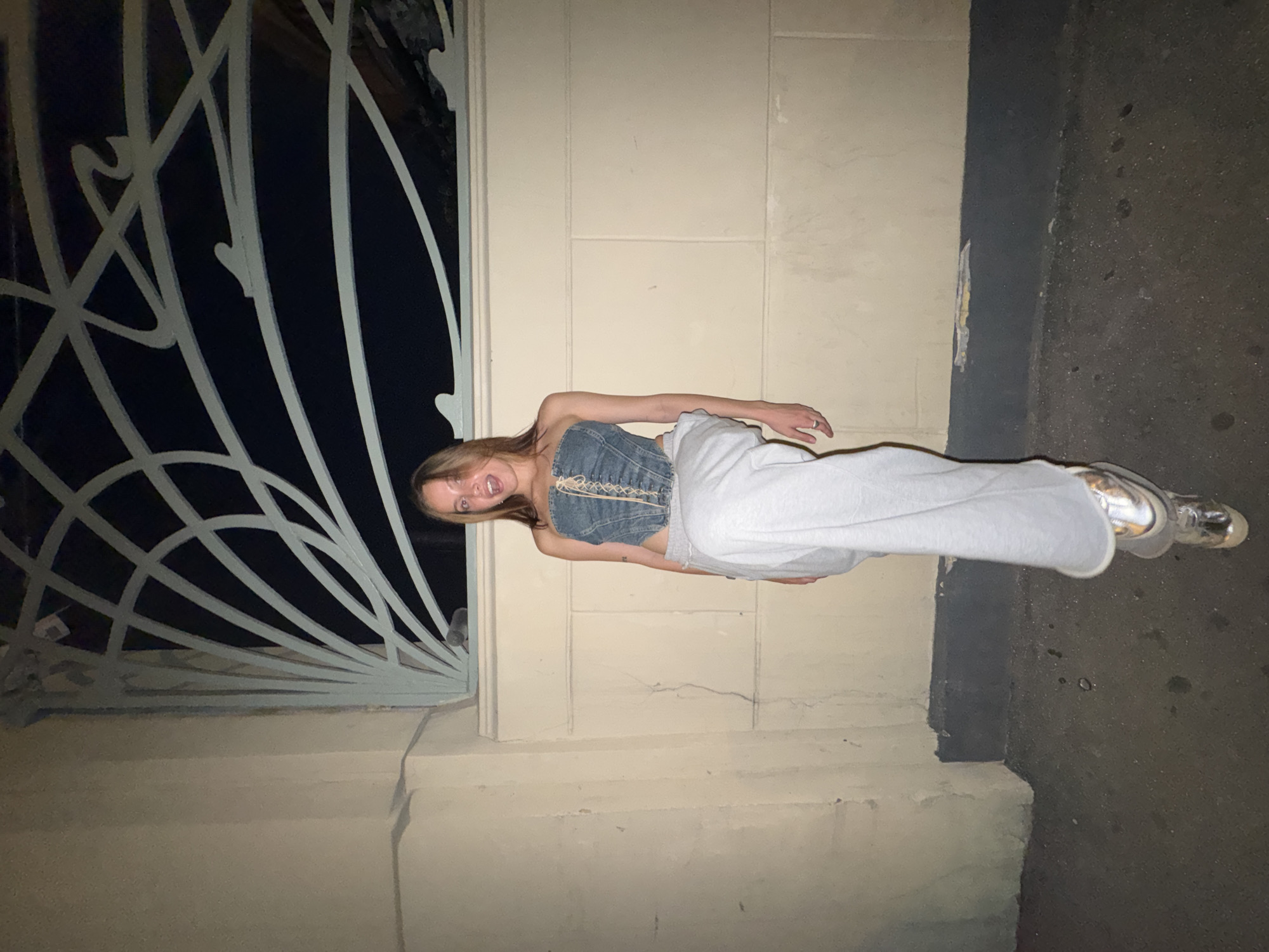 A young woman stands outside a building at night, smiling and looking toward the camera. She wears a denim top and grey sweatpants. The building's exterior features a light-colored wall and an ornate metal gate. The scene appears to be in an urban environment, with pavement visible along the building's base. It is unknown if this image was taken in the city of Укромновское сельское поселение, Ukraine.