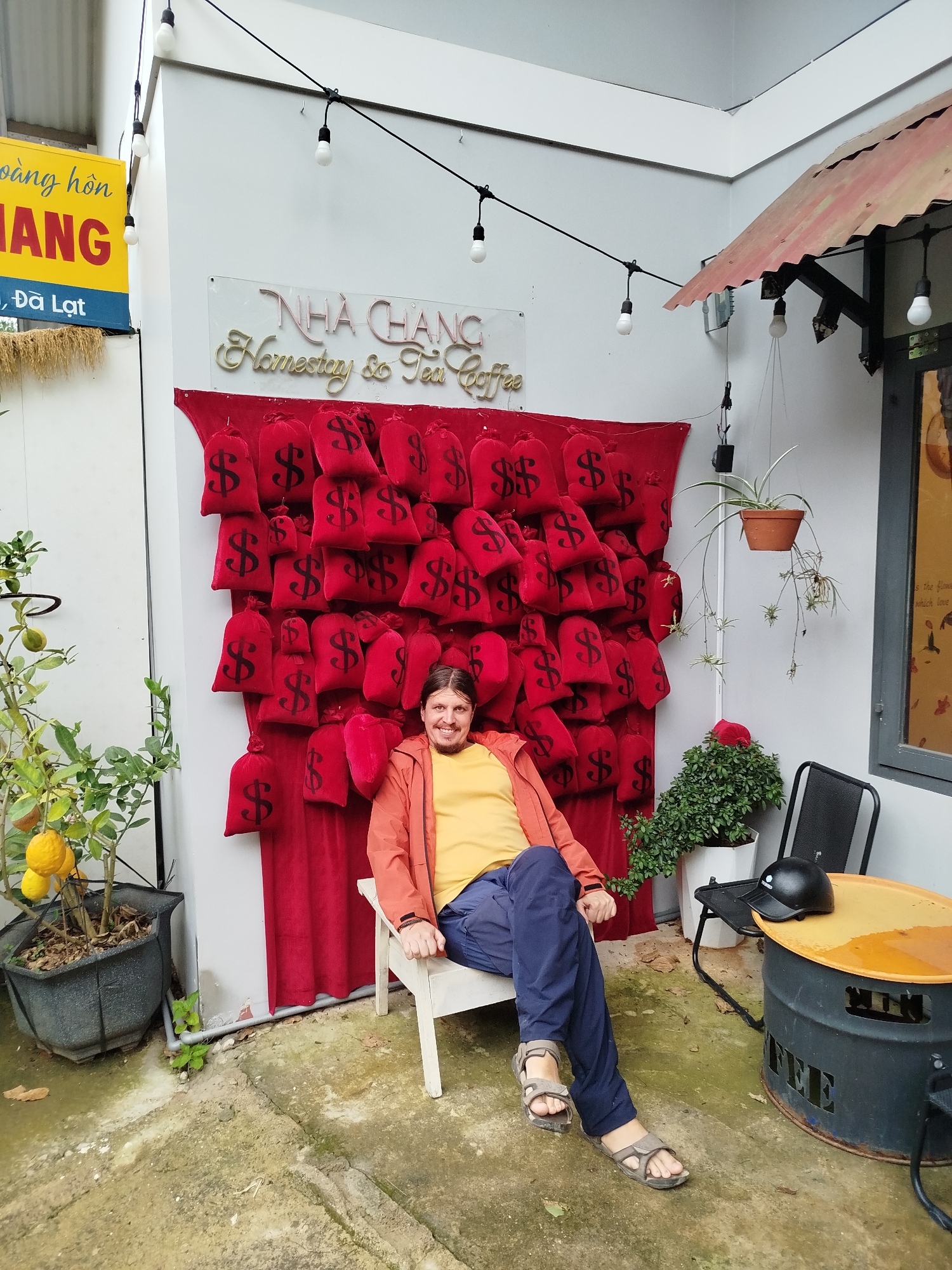 A medium shot captures a middle-aged man seated on a white chair against a wall adorned with red money bags. The man wears an orange jacket over a yellow shirt, along with navy blue pants and sandals. He is smiling towards the camera. Behind him is a red backdrop featuring numerous red bags with a black dollar sign ($) on each.
Above the backdrop is a sign reading "NHÀ CHÀNG Homestay & Tea Coffee." The scene is illuminated by string lights hanging overhead. To the left of the man, a potted lemon tree with ripe lemons is visible. To the right is a green plant in a white pot, a black chair, and a repurposed barrel table with the word "COFFEE" stenciled on it.
In the upper left corner of the image, a sign in Vietnamese, "Hoàng hôn Da Lat", indicates the location as Da Lat, Vietnam. The flooring is concrete, partially covered with what appears to be damp moss or algae, suggesting a humid environment. The architectural setting suggests an outdoor seating area, possibly a courtyard.