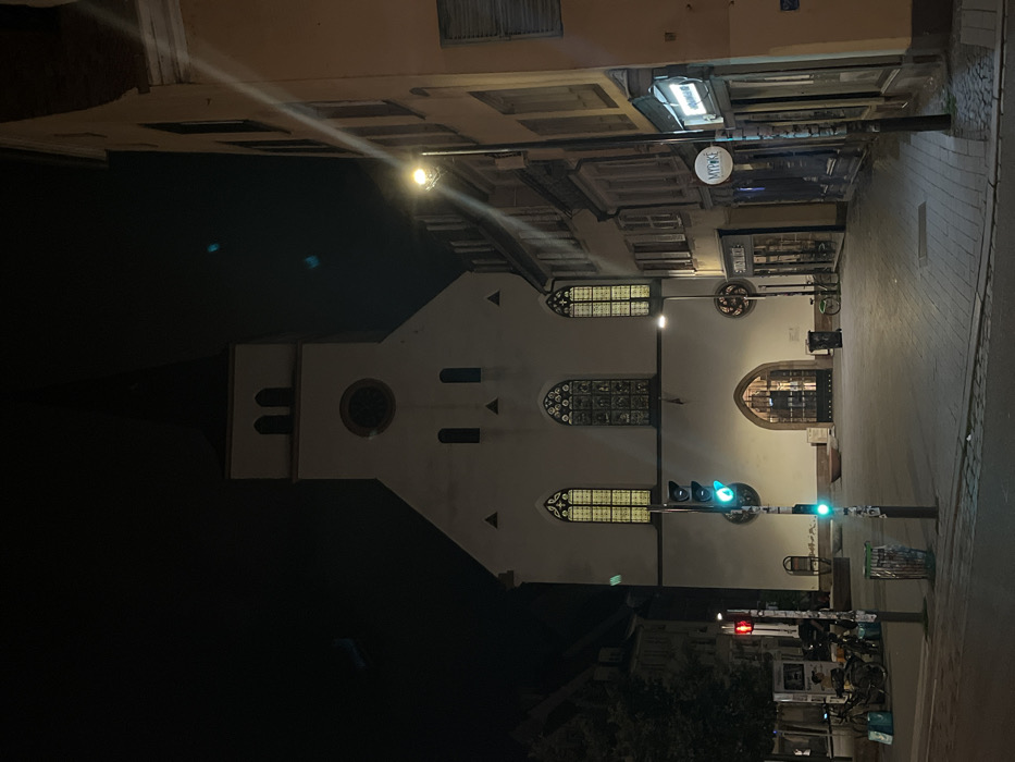 The image displays Église Saint-Thomas (Saint Thomas Church) in Strasbourg, France, during nighttime. The church, a prominent light-colored Romanesque structure with a central spire, is illuminated, showcasing a circular clock face and multiple arched windows, three of which are large and project a yellow glow. Its main arched entrance is gated.
To the left of the church, a paved area contains a traffic light pole displaying a green signal. Further left, a second traffic light shows a red signal. A green cylindrical trash bin is positioned in the foreground. Several bicycles are parked near a low wall or bench, where two indistinct figures are seated.
To the right of the church, a street extends, lined with multi-story buildings. Storefronts are visible along this street, including one with a circular, illuminated sign displaying "MY POKE." Two streetlights, casting bright light and causing lens flares, illuminate this side of the scene. A dark trash bin and a single parked bicycle are also visible near the church entrance on the right. The sky overhead is dark.