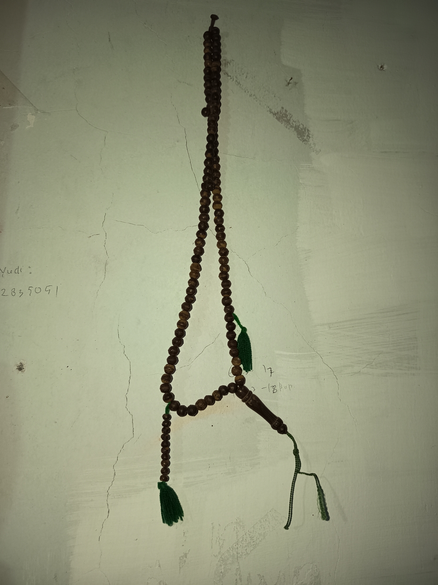 A close-up, medium shot shows a string of prayer beads, made of dark, polished wood, hanging against a pale, cracked, textured wall. The beads are strung on a thin, dark cord. The prayer beads are roughly 70 cm in length and hang vertically. The top of the string is secured to the wall with a small, dark nail. The prayer beads are embellished with two green tassels, one near the top and one near the bottom, and a carved, dark wooden divider. Faint, handwritten marks are visible on the wall to the left of the prayer beads, including what appears to be a name, "Yudi," followed by a number. Additional faint writing is visible to the right of the beads. The lighting is dim and appears to originate from the front, casting subtle shadows. The scene is indoors, in what appears to be a residential or utilitarian space within the city of Krampilan, Indonesia.