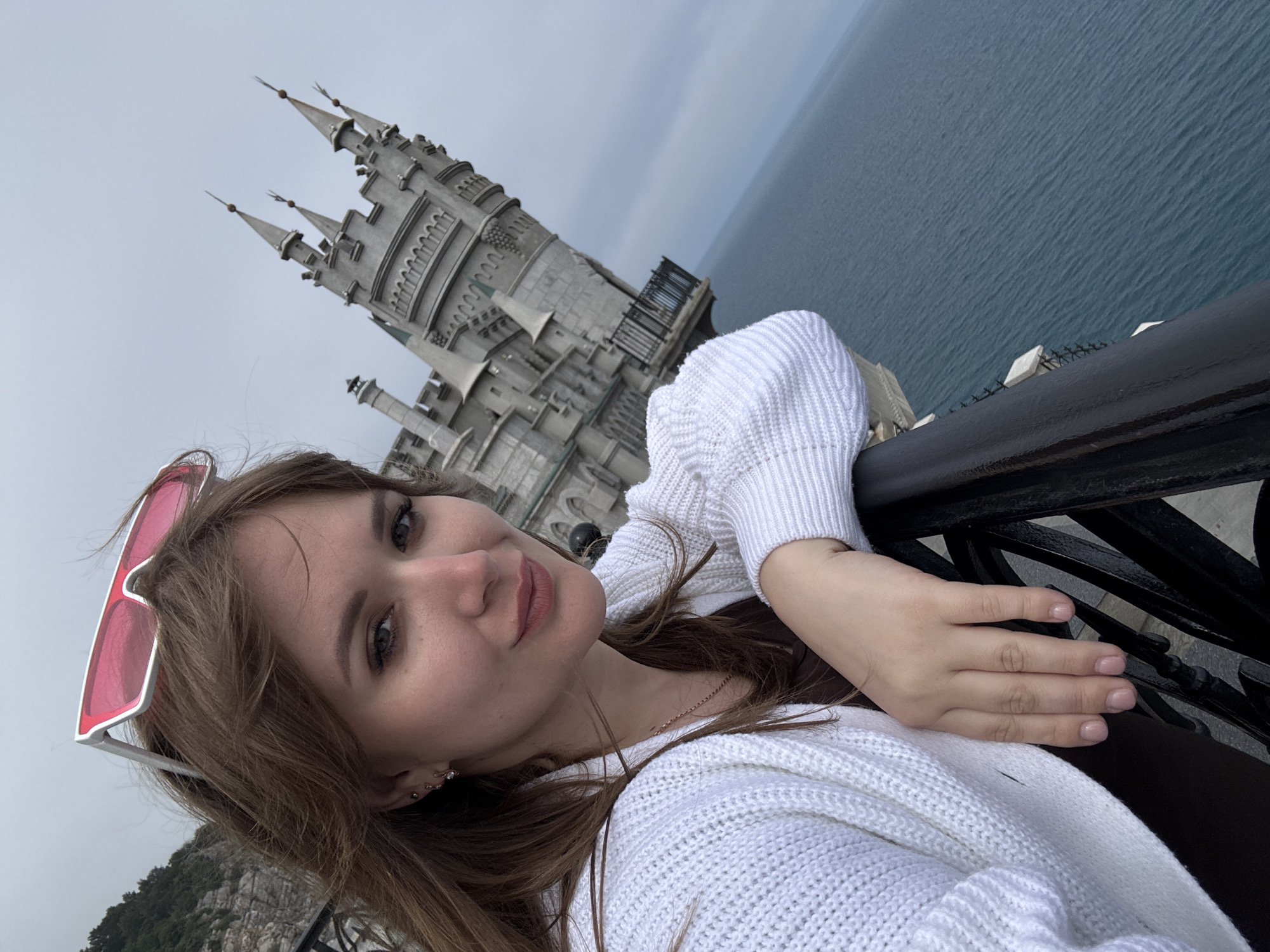 A young woman is taking a selfie with the Swallow's Nest castle in Гаспринский поселковый совет, Ukraine in the background. She is wearing a white cardigan and pink-framed sunglasses resting on her head. She is leaning against a black metal railing. The sea and overcast sky are visible.