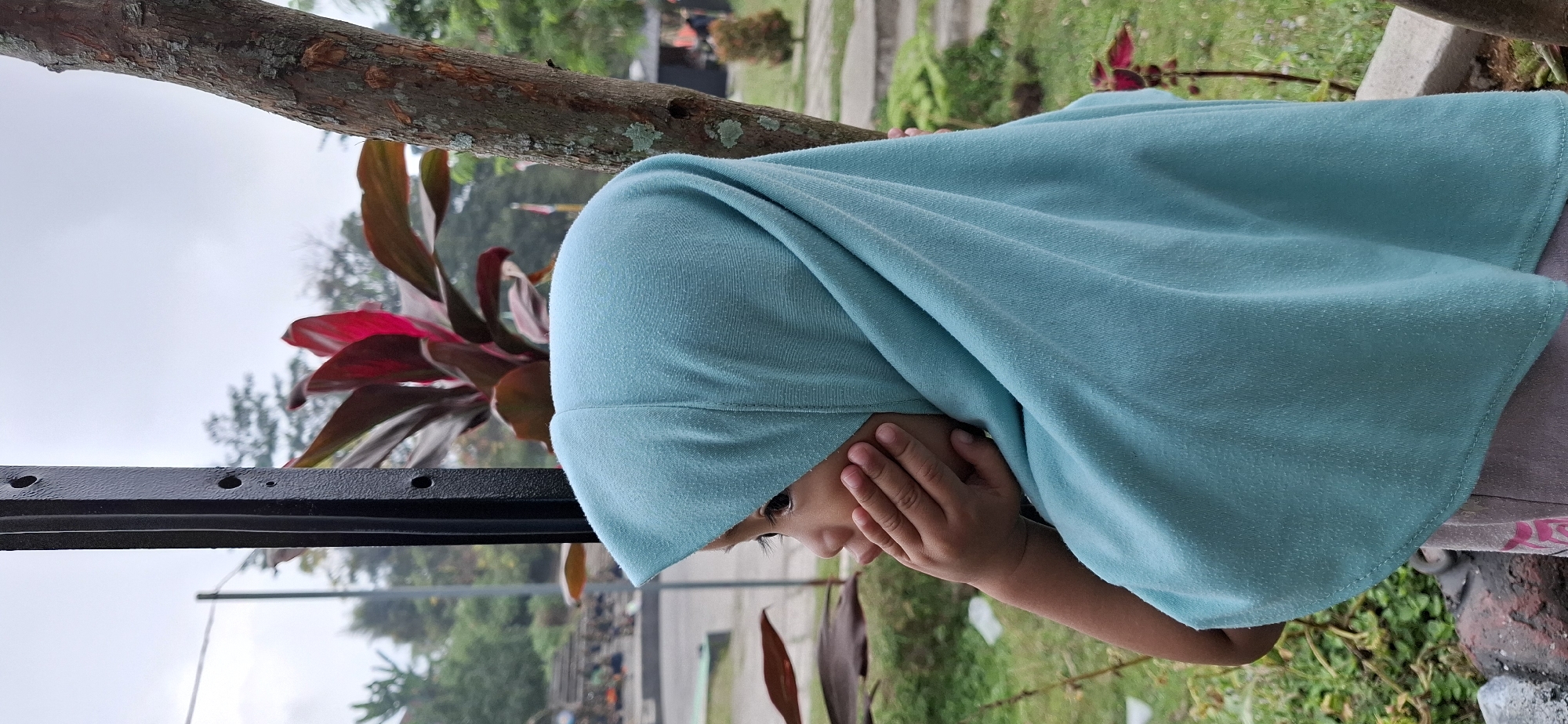 A young girl wearing a turquoise hijab is seen in profile, her right hand resting on her cheek. She appears to be looking off to the left. The background suggests an outdoor setting with lush green foliage, a tree trunk to the right, and a metal pole to the left. The sky is overcast. The scene is likely a public park or garden area in Karang, Indonesia.