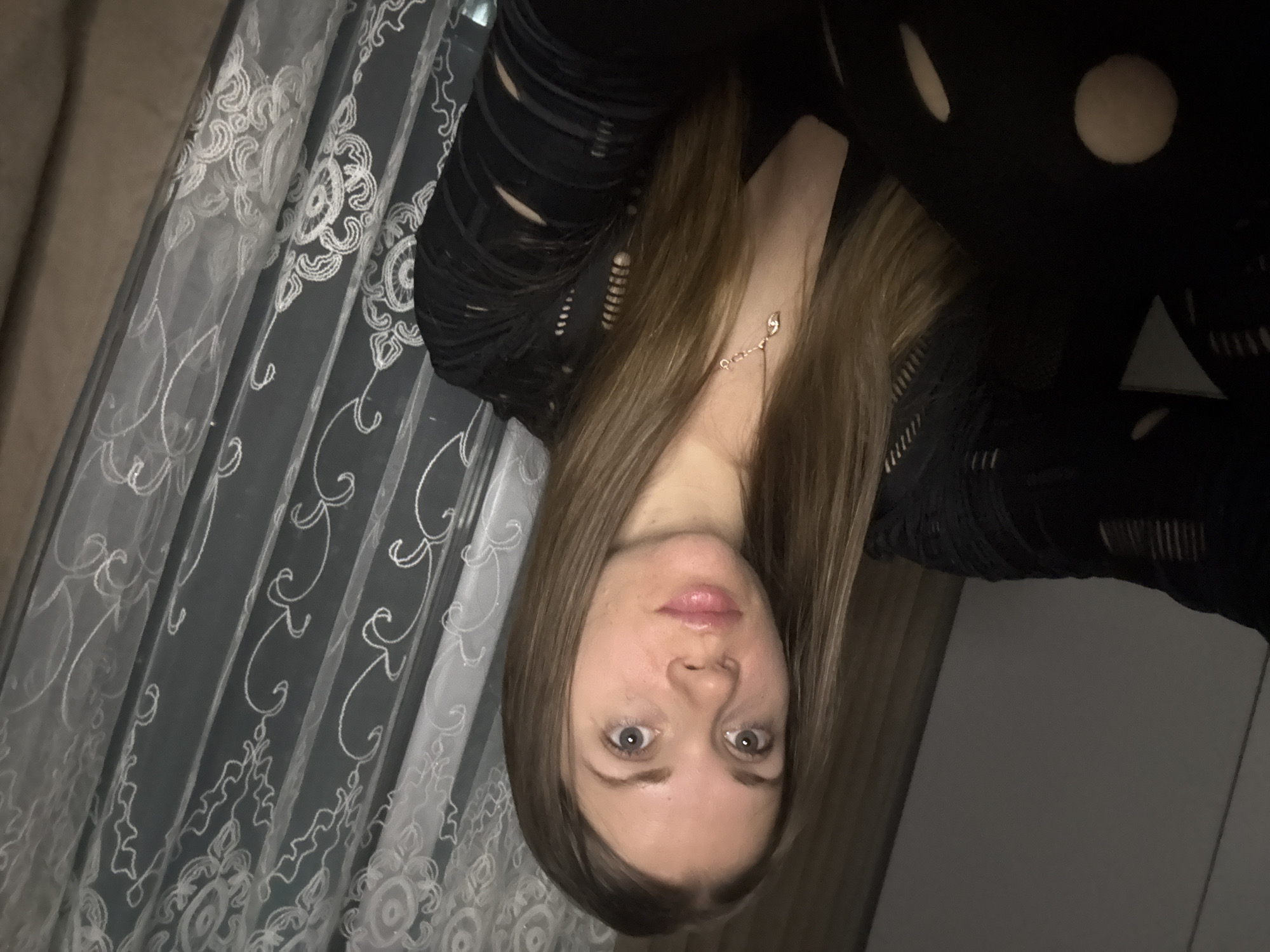 A woman with long brown hair, fair skin, and light eyes is centered in the image, appearing to look directly at the camera. She wears a dark top with patterned or distressed details. The background includes a sheer curtain with an ornate pattern on the left side and a solid gray or dark background on the right, suggesting an indoor setting. There are no details in the image that would suggest the location is Batumi, Georgia.