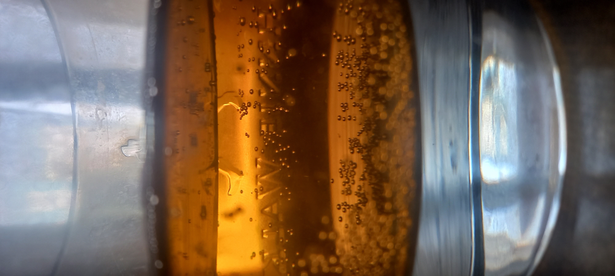The image shows a close-up of what appears to be a liquid, possibly beer or a similar beverage, contained within a glass or transparent vessel. Bubbles are visible throughout the liquid. The vessel has a rounded, possibly bulbous shape. Part of the vessel is clear, while another section has a brownish-amber hue. Given the level of detail, the photo might be a macro shot, capturing minute details of the bubbles and the texture of the glass. There is no information in the image to connect it to a specific location.