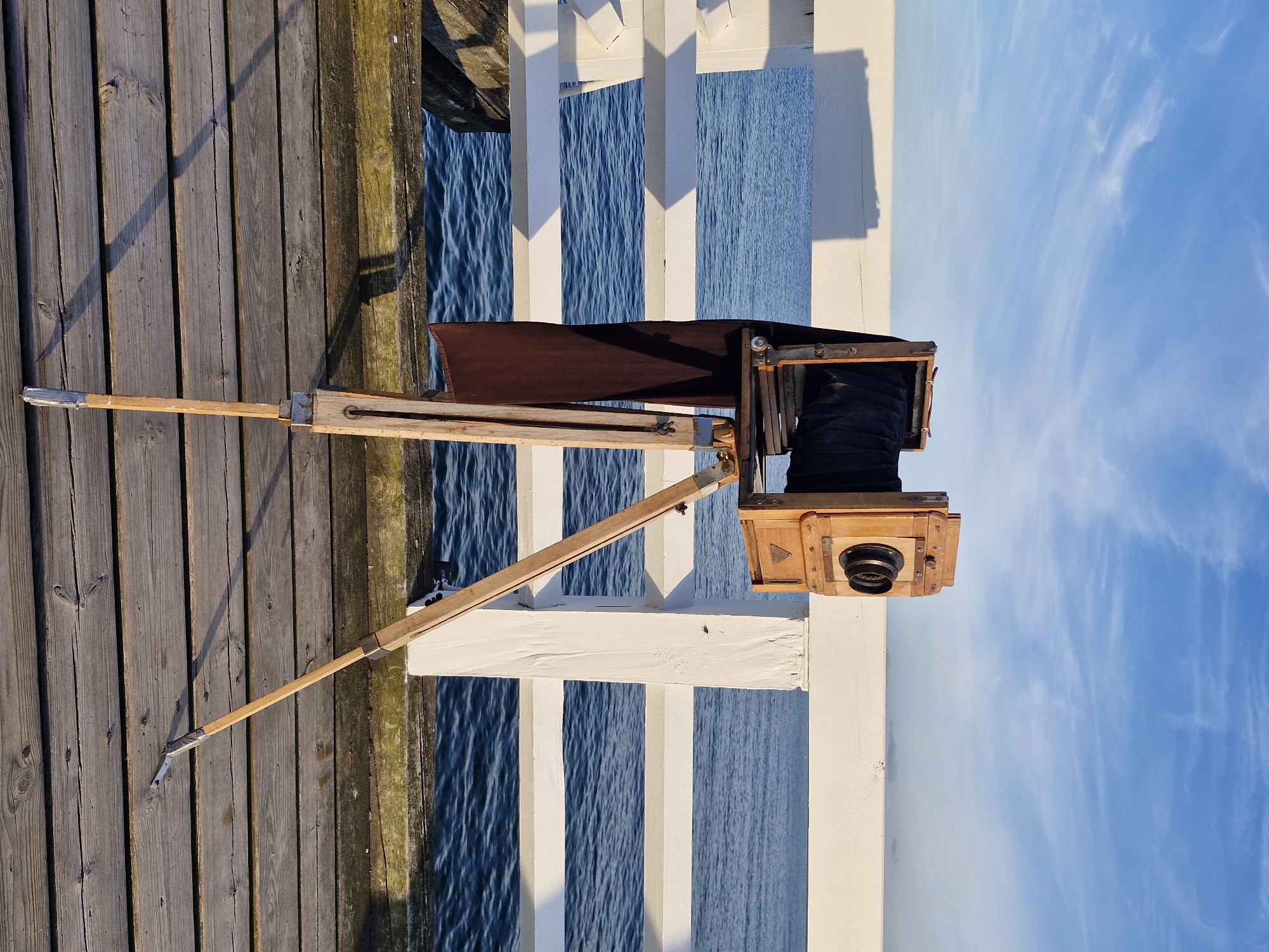 An antique wooden camera sits on a tripod on a wooden pier in Sopot, Poland. The camera is positioned facing the sea, with its bellows extended. The pier's wooden planks are worn and weathered, and a white railing runs along its edge. The sea is calm and blue, and the sky above is a clear, bright blue with a few wispy clouds. The scene is bathed in the warm light of what appears to be late afternoon or early morning. The camera and tripod are the sole subjects of the image, with no people present. The focus is on the vintage equipment against the backdrop of the natural environment.