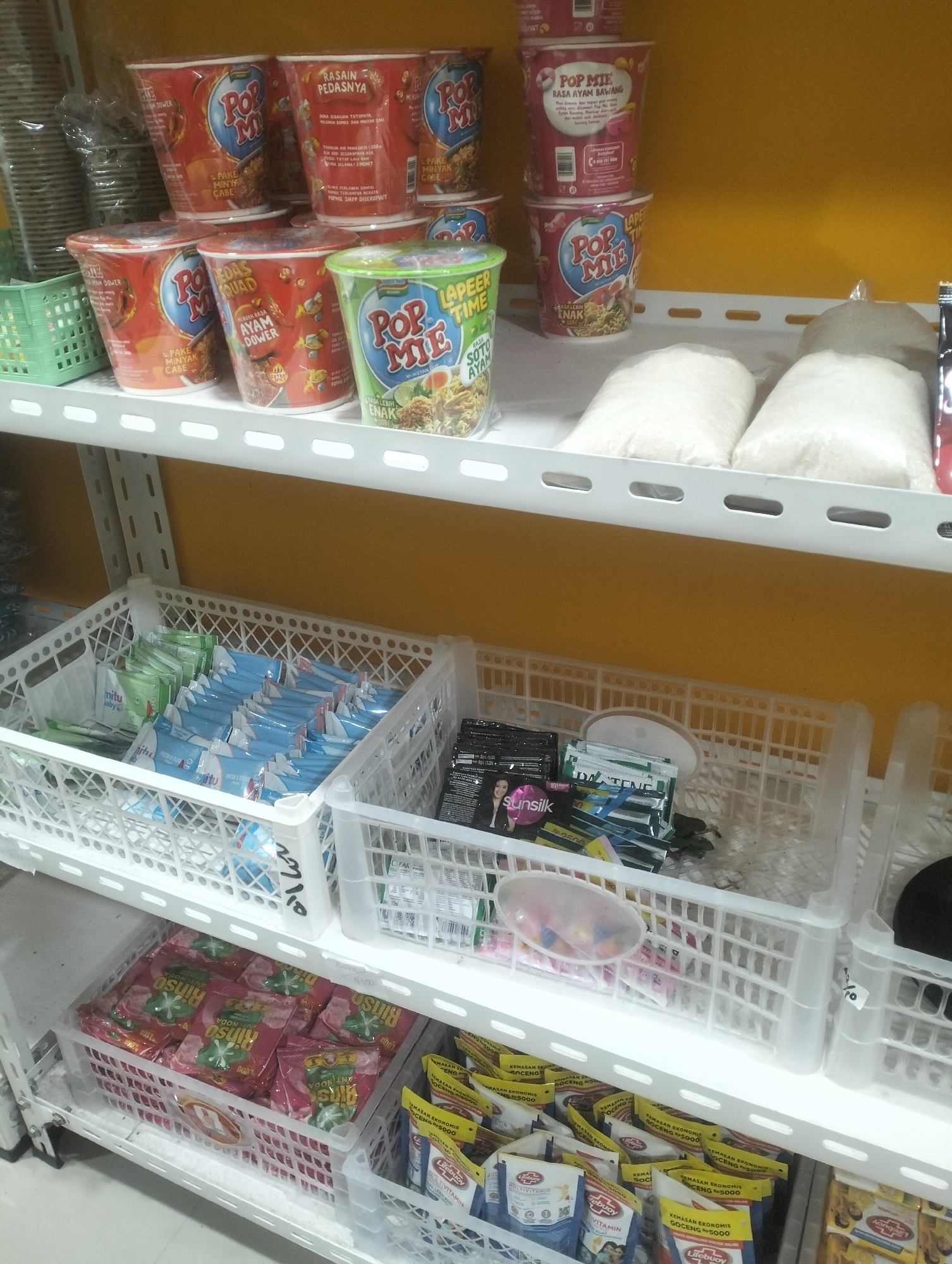 The image depicts a retail store shelf in Air Jamban, Indonesia, stocked with various consumer goods. On the top shelf, several red and green cup noodles are stacked, predominantly of the "Pop Mie" brand. Below this, the middle shelf displays two large white bags, likely containing rice or sugar, and several white plastic baskets filled with smaller packaged goods such as shampoo sachets, detergent packets, and possibly other personal care items. The bottom shelf is also lined with white plastic baskets containing more packaged goods, including laundry detergent packets with the "Rinso" brand and sachets of "Lifebuoy" multi-vitamin. The overall environment suggests a well-organized retail display within a local Indonesian store.