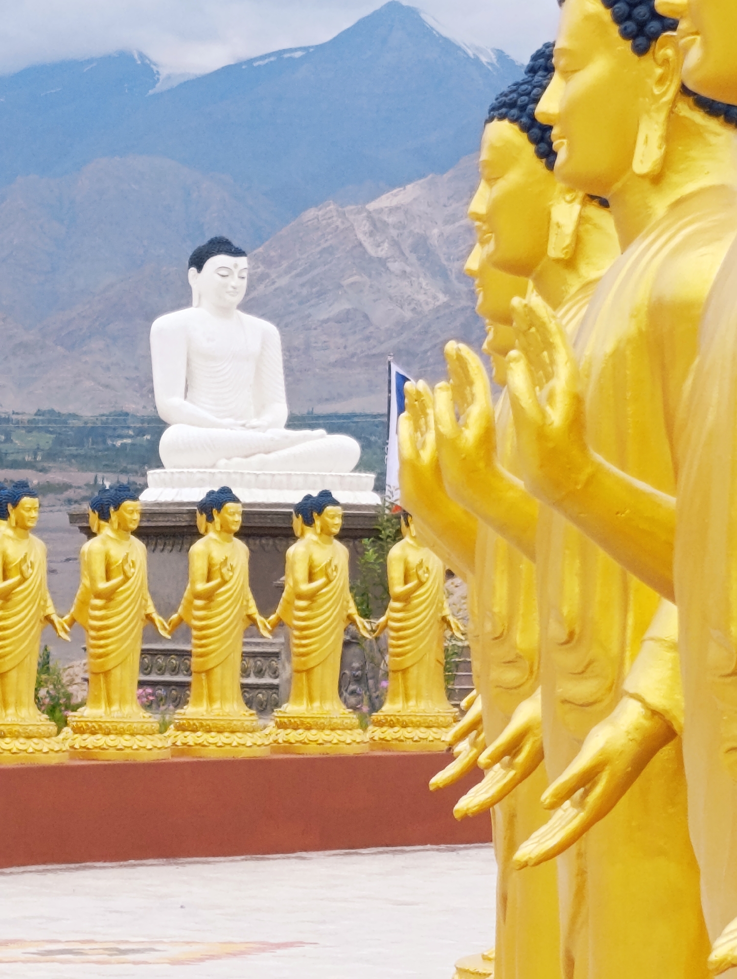 This is a photograph of the Maitreya Buddha statue complex in Leh, India. In the foreground, a row of golden Buddha statues stand in a line, facing to the left of the frame. Their hands are raised in a gesture of blessing or prayer. Behind these golden statues, a larger, white Buddha statue is seated in a meditative pose on a raised platform. In the background, rugged mountains rise, their peaks capped with snow under a cloudy sky. The scene appears to be an outdoor temple or monastery courtyard.