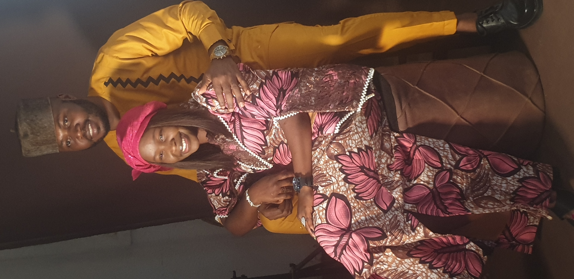 A portrait photograph captures a smiling man and woman standing close together indoors, presented as if rotated 90 degrees clockwise.
The man, positioned slightly behind and to the left of the woman, wears a mustard yellow long-sleeved top featuring a dark zig-zag pattern across the chest, and matching trousers. He sports a dark, traditional cap (fila) and a gold-toned watch on his left wrist. With a broad smile, he looks directly at the camera, his left arm gently around the woman's waist.
The woman, standing in front, wears a vibrant, long-sleeved dress or wrapper made from patterned fabric in shades of pink, brown, and white, adorned with large floral designs. The garment features intricate white beaded or embroidered trim along the neckline and cuffs. She dons a bright pink headwrap (gele) and dark, flowing hair, smiling broadly towards the camera. She wears a dark watch on her left wrist, a silver-toned bracelet on her right, and a ring on her right hand.
Both individuals convey expressions of joy and affection. The background is a mix of a dark, plain wall behind the man and a rich brown, textured piece of furniture, possibly a sofa or chair, behind and beside the woman. The attire worn by the couple is indicative of traditional Nigerian fashion. The scene is softly lit, suggesting an indoor setting. There is no visible text in the image.