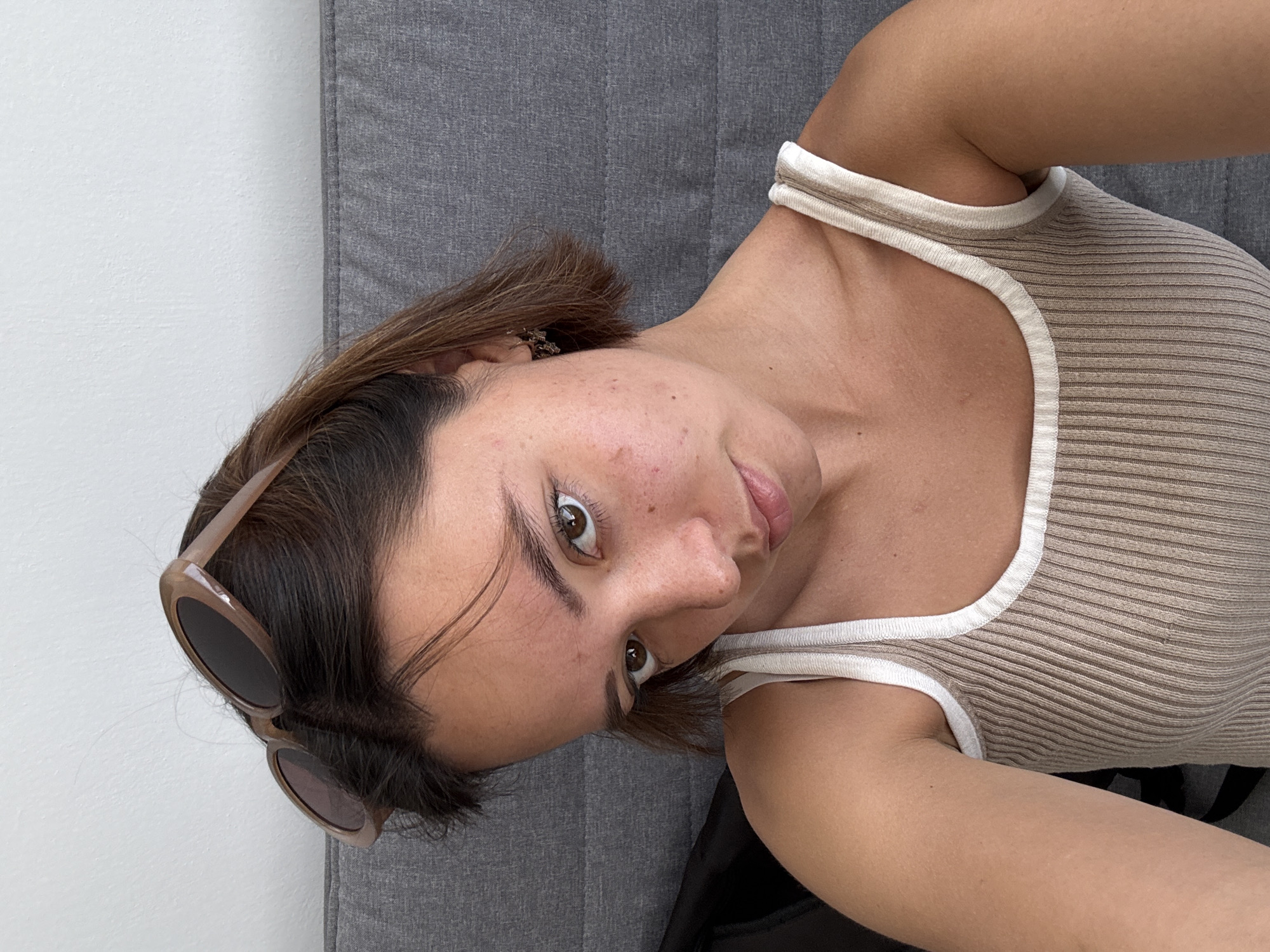 A woman with short brown hair, light skin, and brown eyes is pictured. She is wearing a beige and white tank top. Sunglasses are visible on the left side of her head. She is lying on a gray surface. The location is unknown, and there is no evidence that it is in San Bernardino Tlaxcalancingo, Mexico.