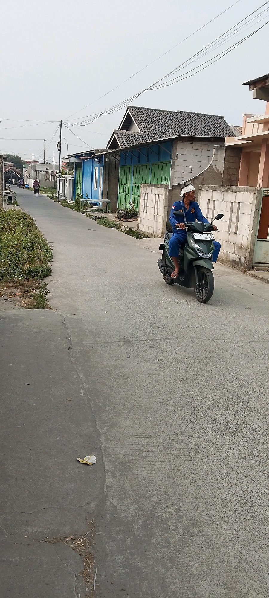 An elderly man wearing a blue uniform and a white headscarf rides a dark green scooter down a paved road in a residential area. The scooter has a license plate that reads "B 6227 YED". The man is looking to his right, away from the camera. The road is flanked by houses and low walls. To the left of the road, there is overgrown vegetation. In the background, other houses and a utility pole with wires are visible. The sky is overcast. The scene appears to be in Sriamur, Indonesia.