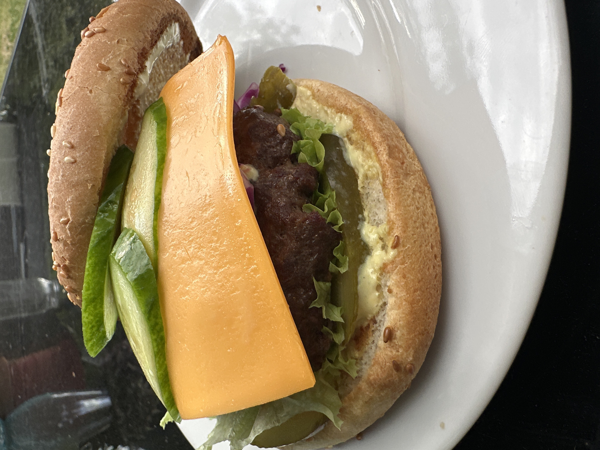 A close-up, high-angle shot shows a cheeseburger on a white plate. The burger is assembled on a sesame seed bun, with a thick beef patty topped with a slice of bright orange cheese. Additional layers include lettuce, pickles, and cucumber slices. The background is dark and blurred, suggesting an outdoor setting with a black, reflective surface, possibly a table. The scene is set in Krefeld, Germany.