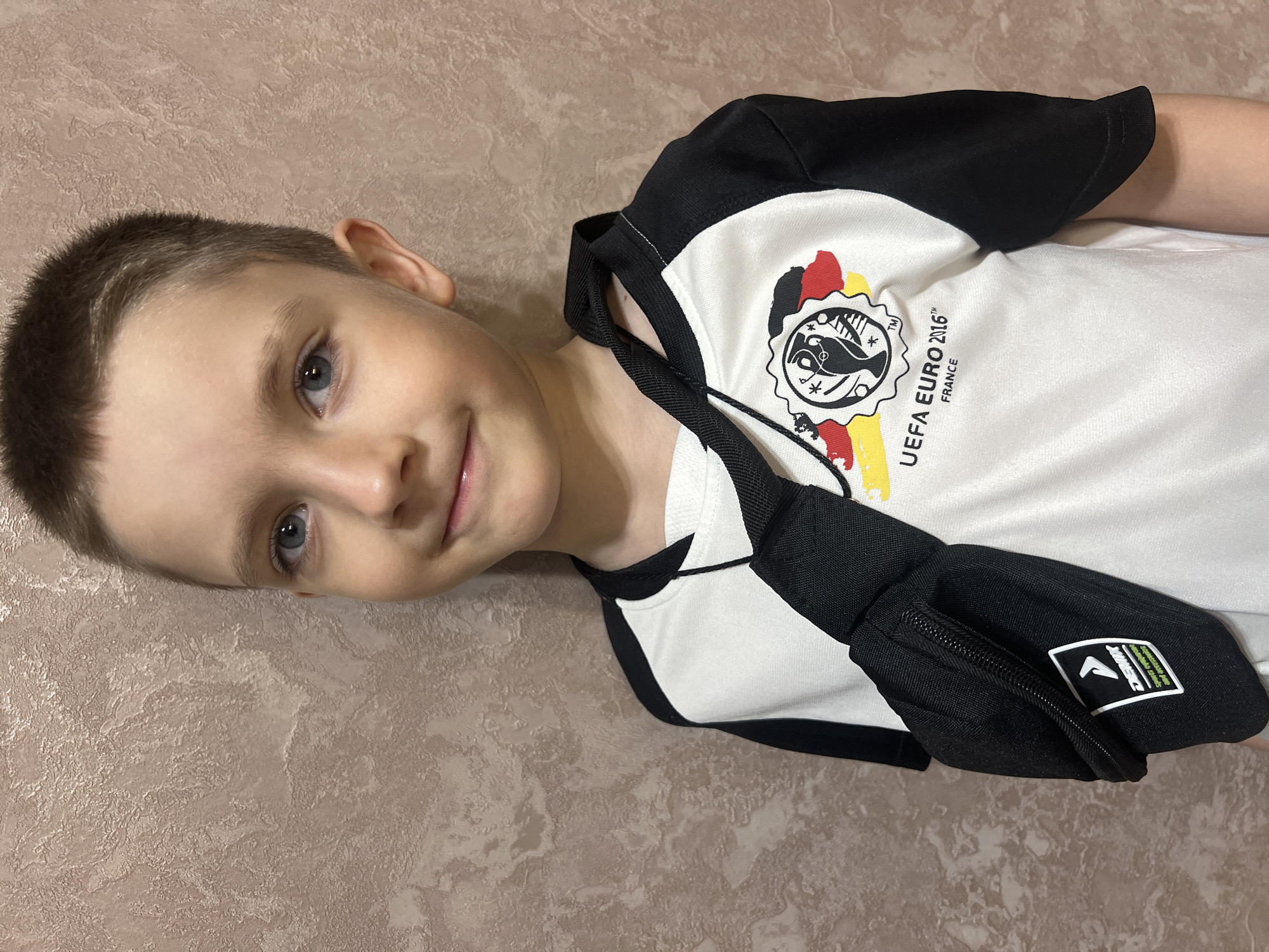A young Caucasian boy with short brown hair and blue eyes smiles slightly, facing the viewer while slightly tilted to the left. He is wearing a white and black t-shirt with a logo that includes the words "UEFA EURO 2016™ FRANCE". He is also wearing a black fanny pack slung across his body. The background is a textured wall with a beige or light brown color. The photo was taken in Марковский, Russia.