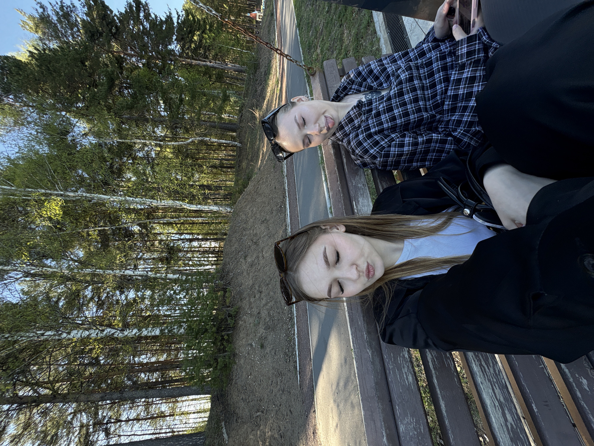 Two young women are lying on a wooden park bench outdoors. The bench is situated along a paved pathway that runs through a forest of tall, slender trees. One woman is wearing a black and white plaid shirt and has sunglasses perched on her head. She is holding a cell phone. The other woman is wearing a light blue shirt under a black jacket and also has sunglasses on her head. The location is in the city of Chaikovsky, Russia.
